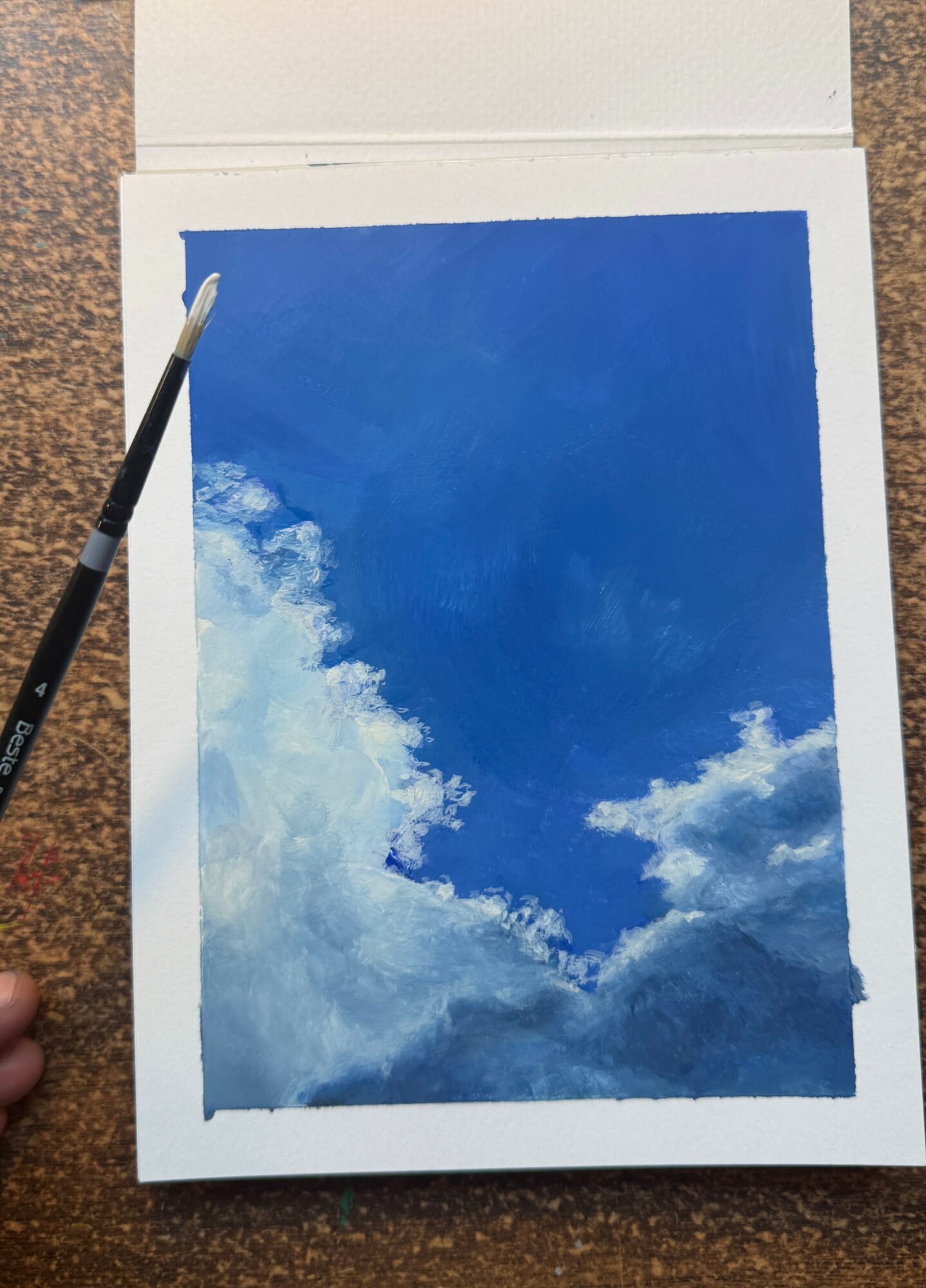 Painting of a blue sky with white clouds, on paper with a paintbrush nearby.