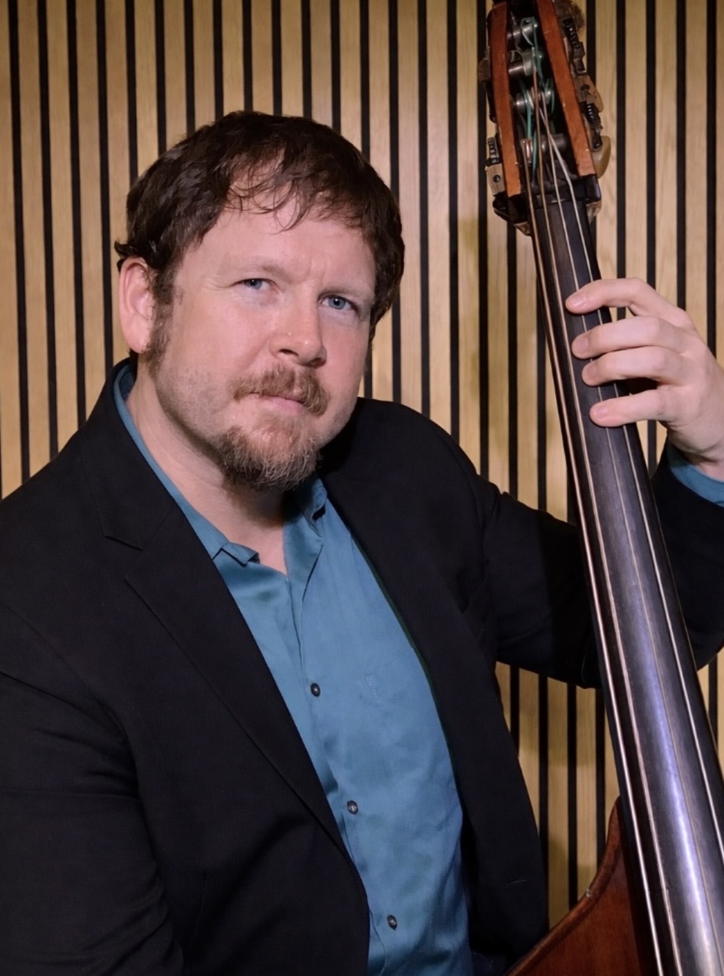 Man with beard and mustache in a black blazer and blue shirt holding a double bass, standing in front of a wooden panel background.