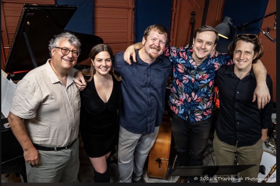 Group of five people smiling and posing together in a music studio, with musical instruments and equipment in the background.