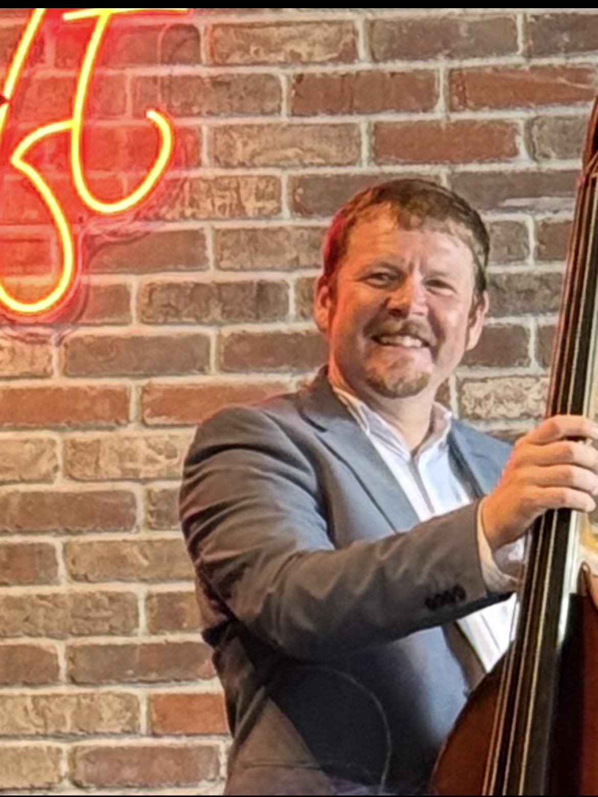 Man in a suit playing a double bass against a brick wall with a neon sign, smiling.