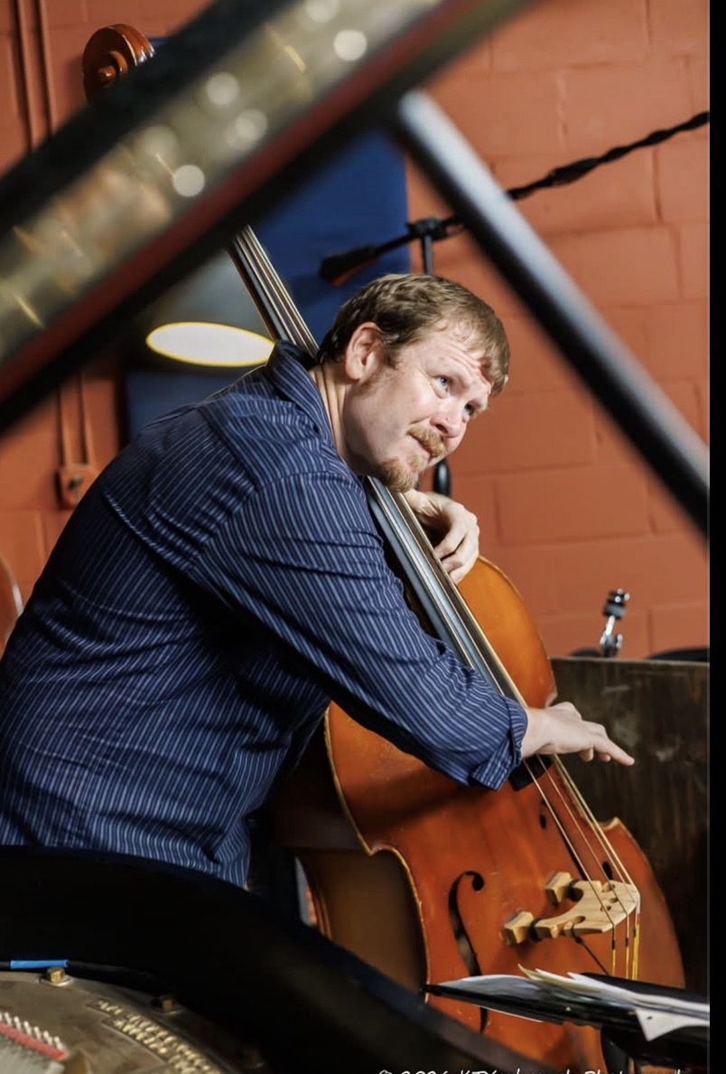 Man playing a cello, leaning on a piano, in a room with brick wall background.