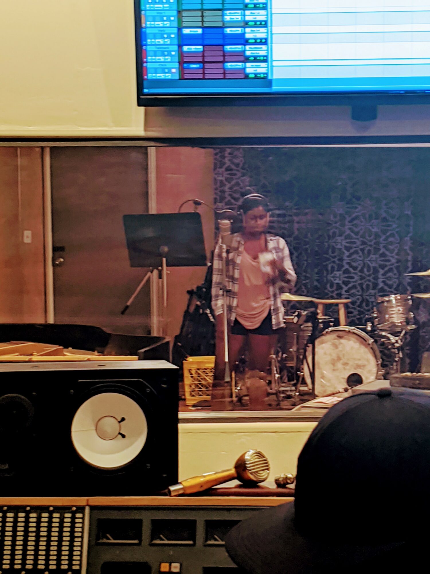 Person standing in a music studio with drums and equipment, reflected in a glass window.