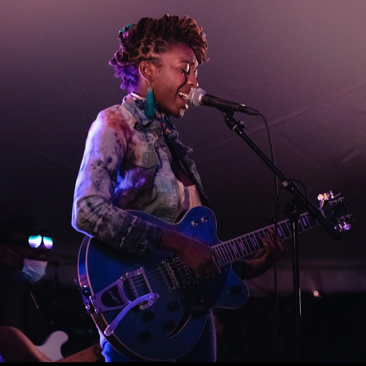 Person singing into a microphone while playing an electric guitar on stage, with purple lighting in the background.