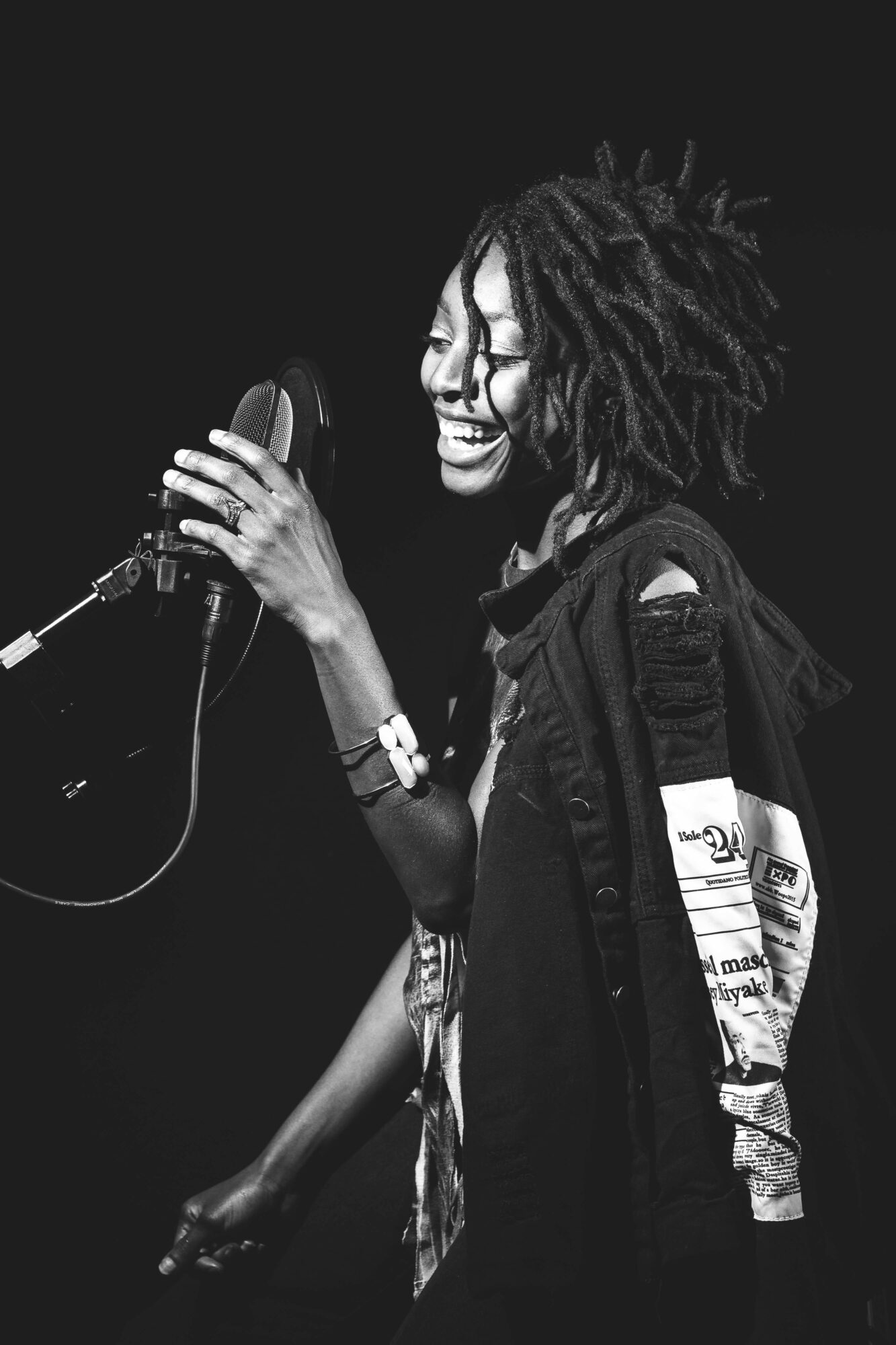 Woman with dreadlocks singing into a microphone, smiling, wearing a jacket with patches, in a dark setting.