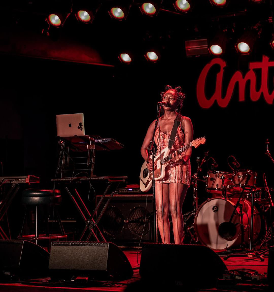 Musician performing on stage with guitar, microphone, and drum set, illuminated by stage lights, with red background and partially visible word 'Am'.