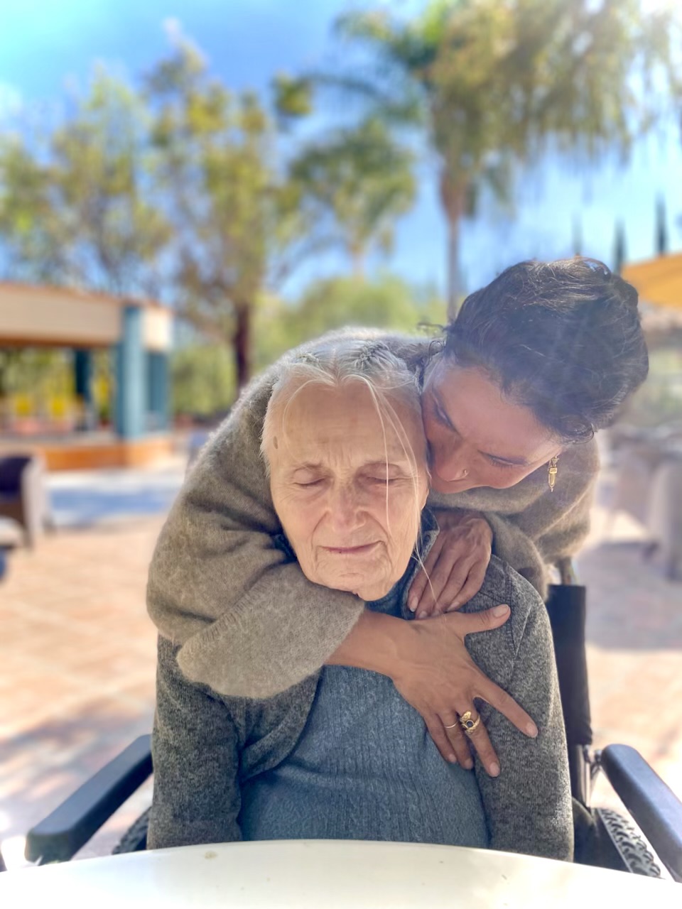 A young person hugging an elderly woman outdoors, with trees and a building in the background.