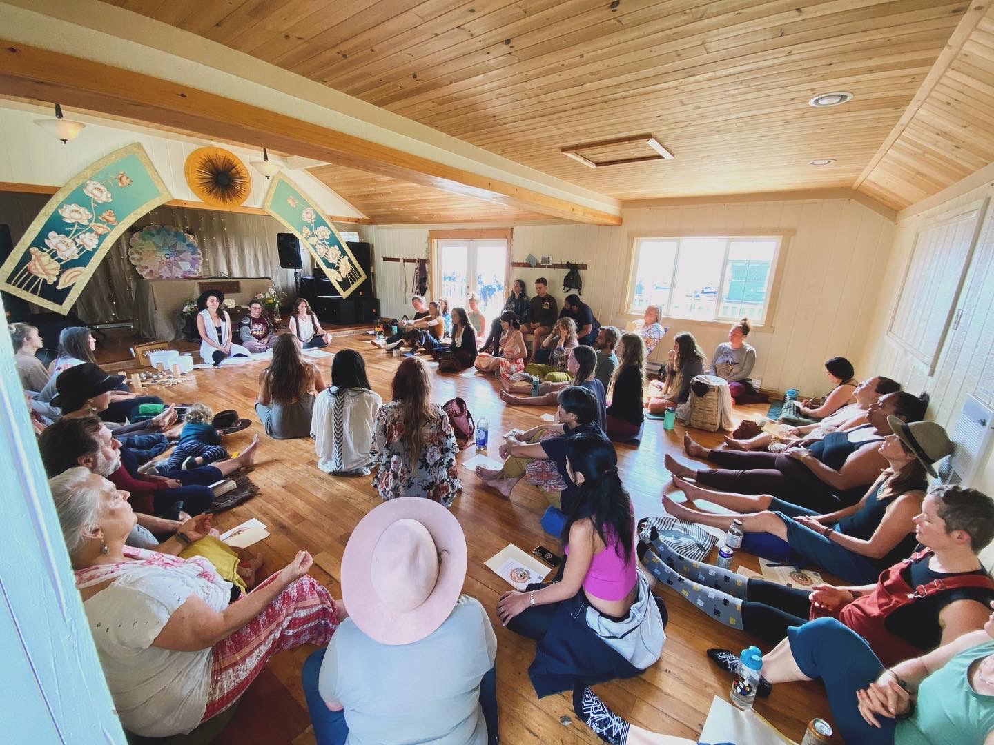 Group of children and adults seated on floor in a room with wooden ceiling, watching a performance on stage with decorations.