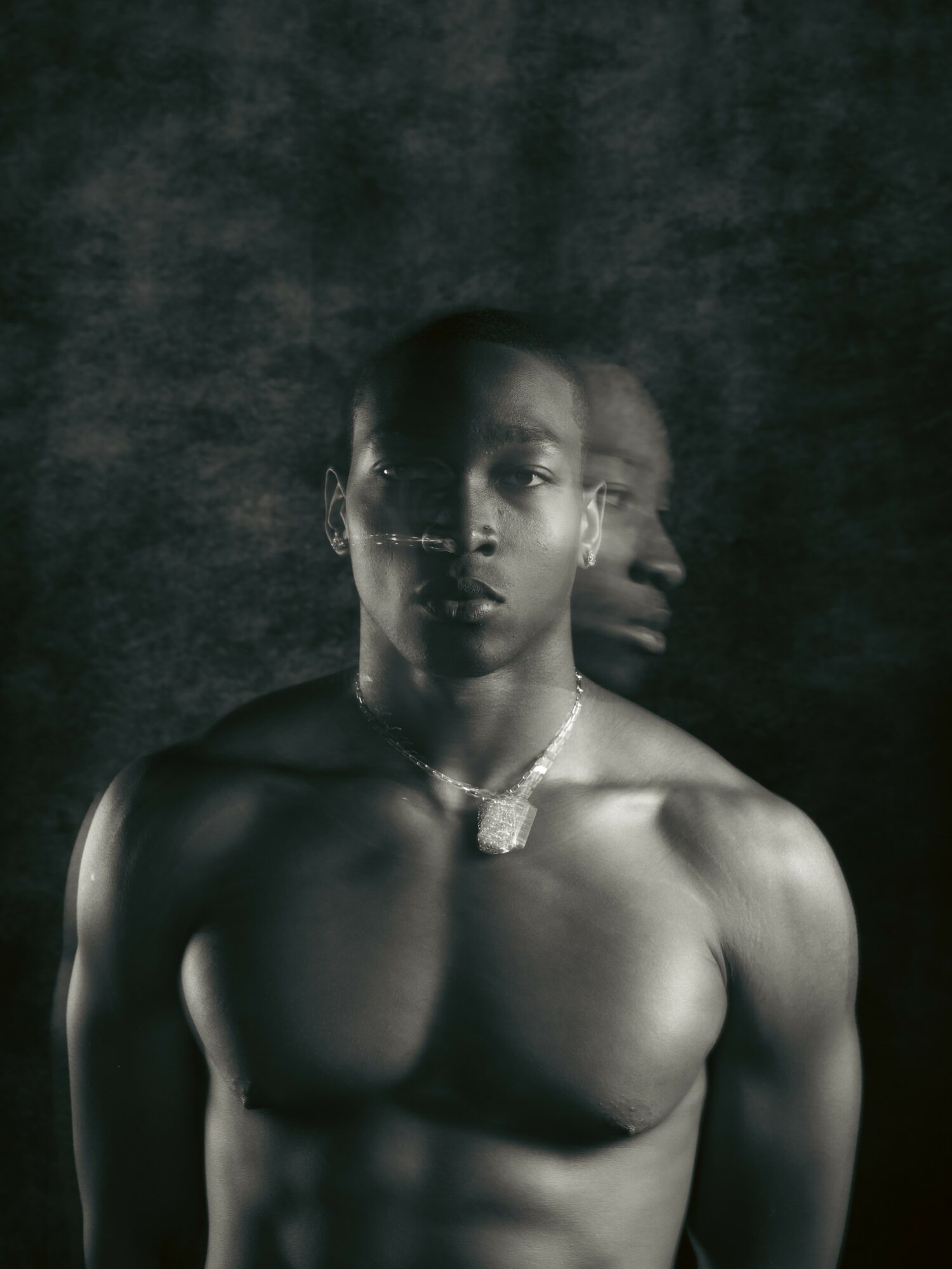 Black and white portrait of a shirtless person with a necklace, facing forward with a dark textured background.