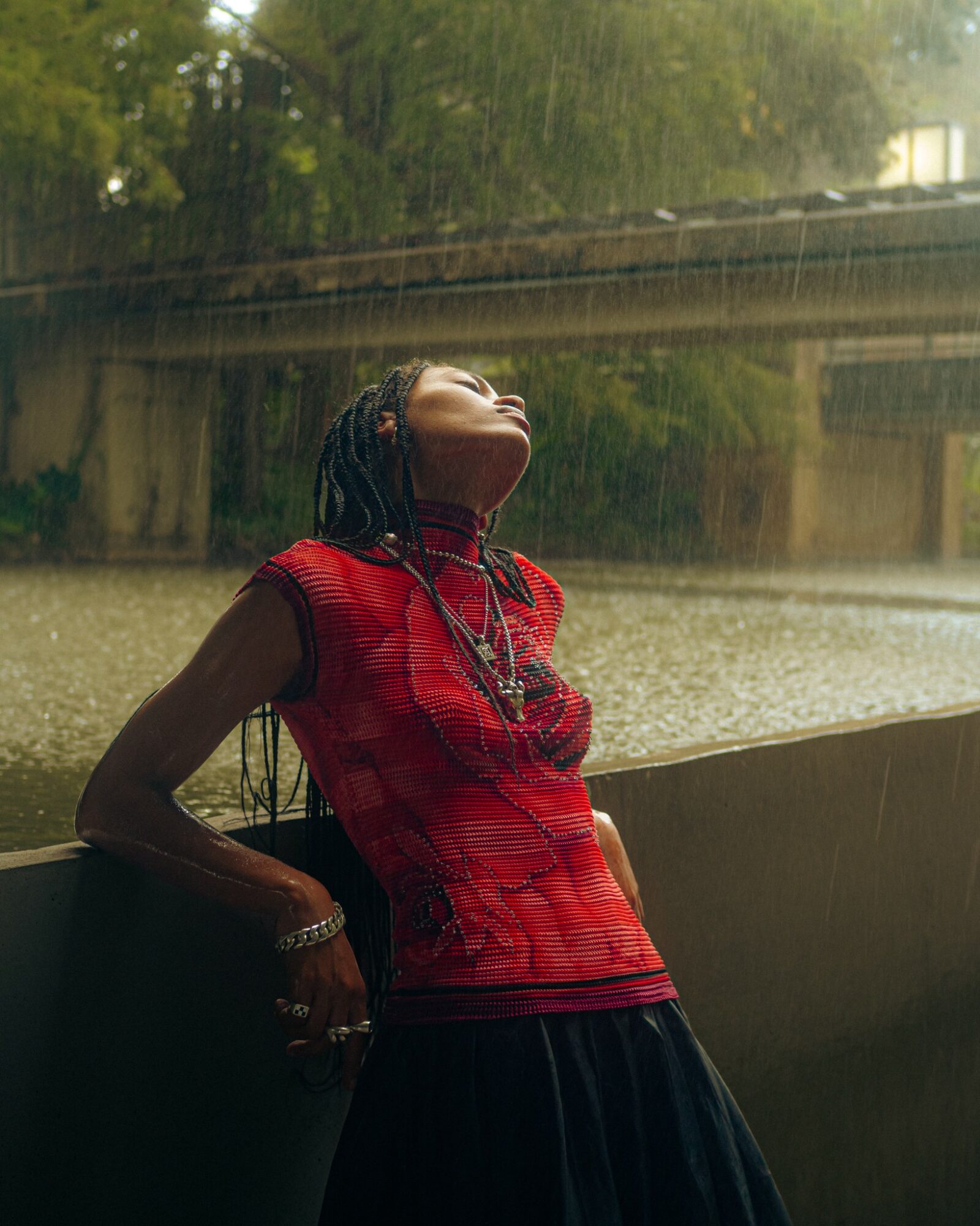 Person with braided hair wearing a red top and black skirt, leaning against a window during rain, looking upward.