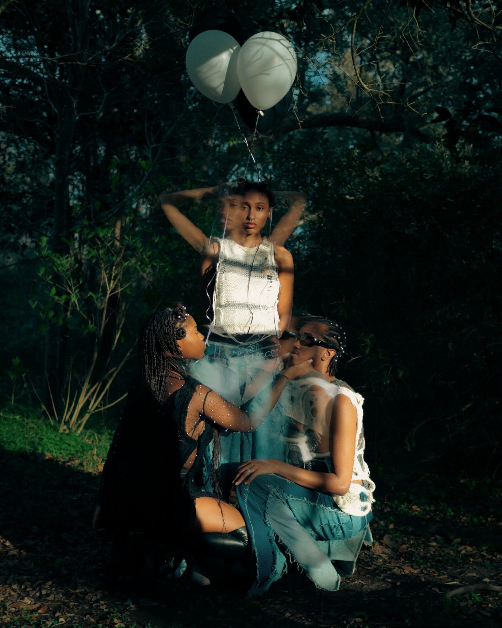 Three people outdoors at night, one standing with balloons, two sitting and talking, surrounded by trees and foliage.