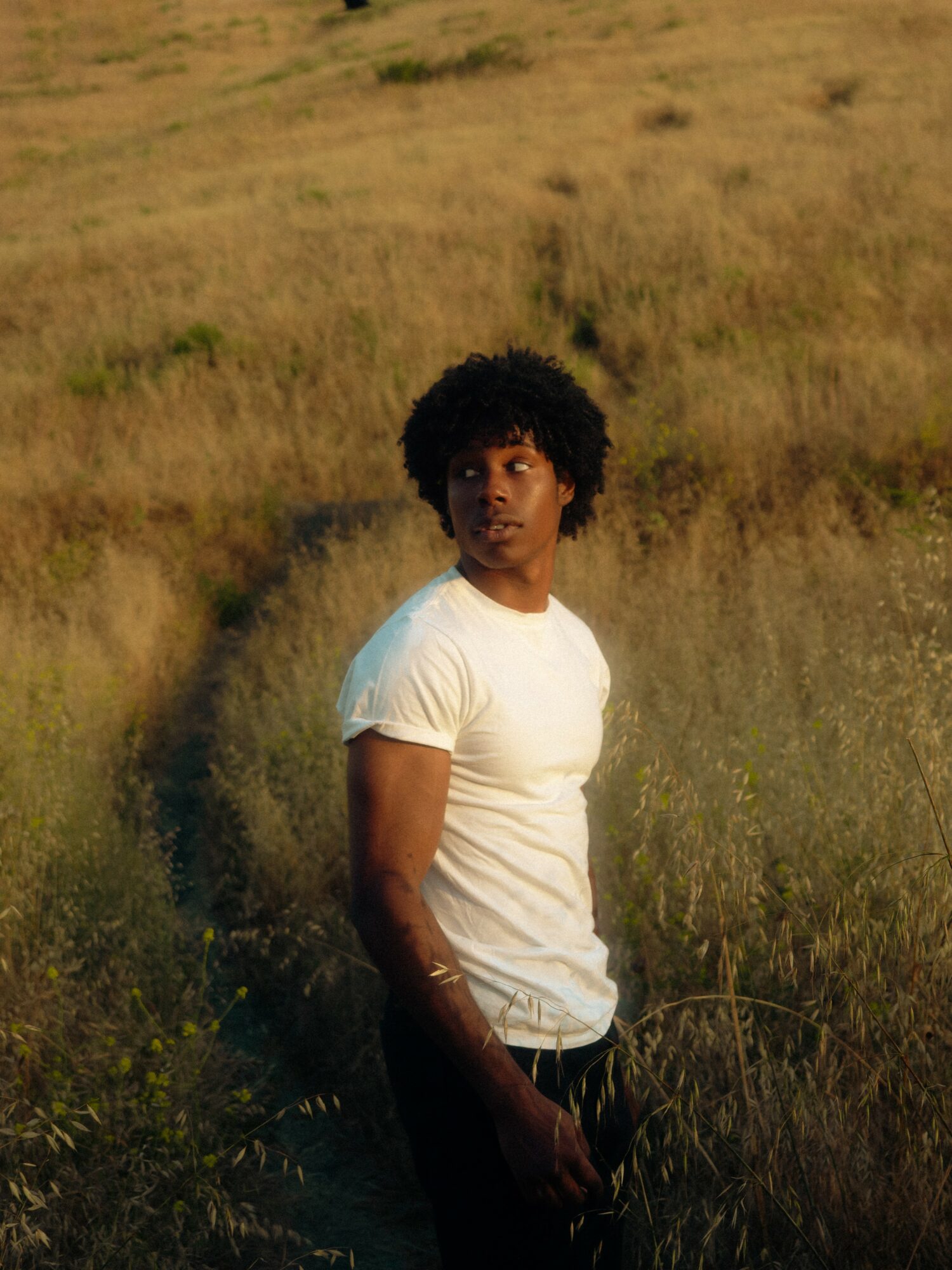 Young person with curly hair in a white t-shirt standing in a grassy field during daytime.