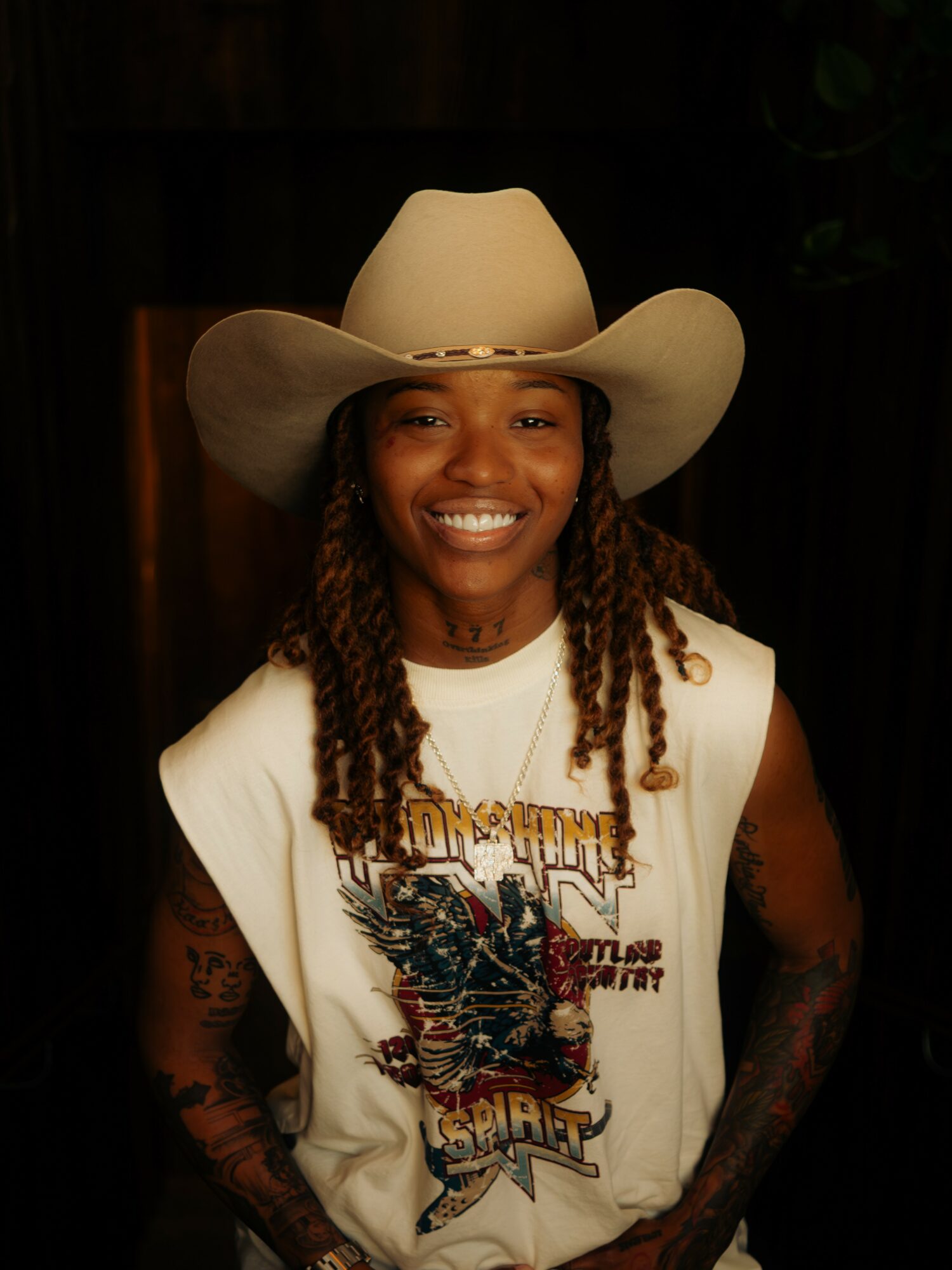 Smiling person wearing a large cowboy hat and a sleeveless graphic T-shirt, standing indoors with dark background.