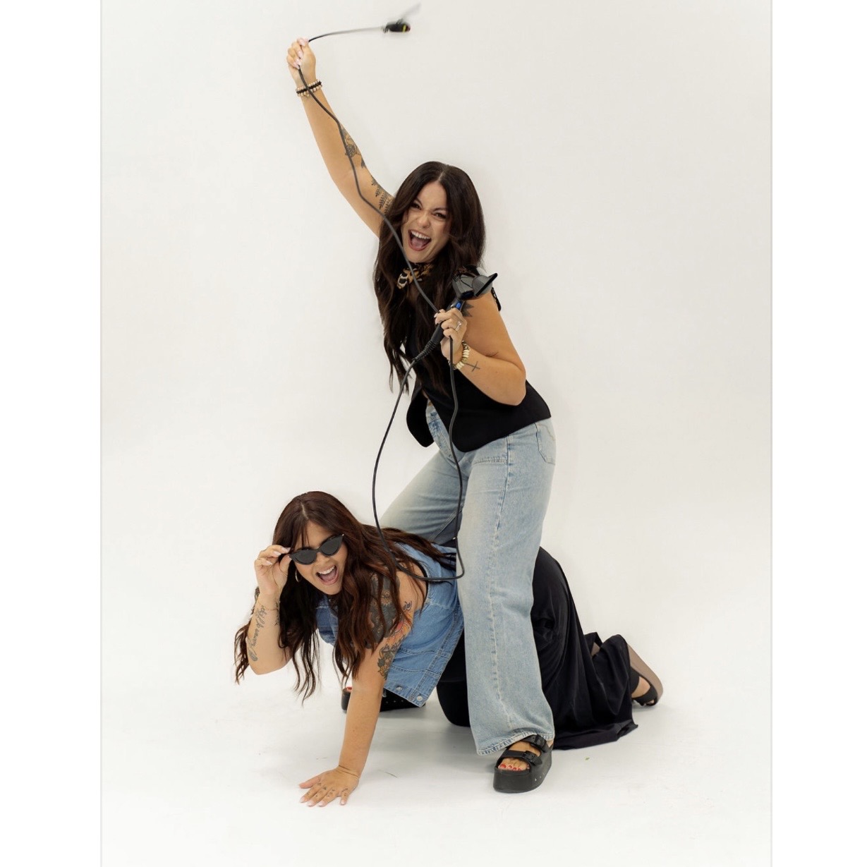 Two women posing playfully, one standing and holding a whip, the other on hands and knees, both smiling, against a white background.