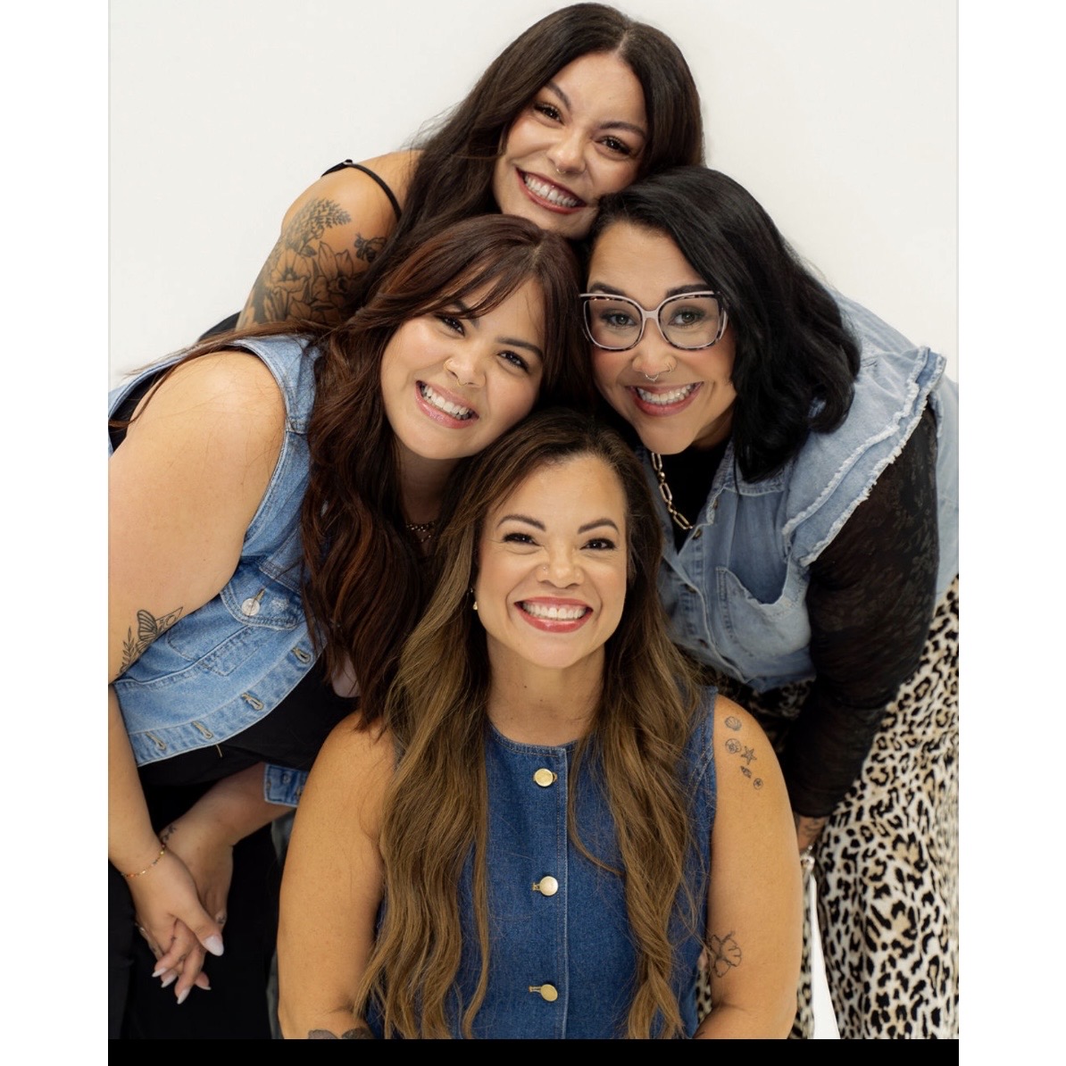 Four women smiling and posing together, with one woman in front and three behind, against a plain background.