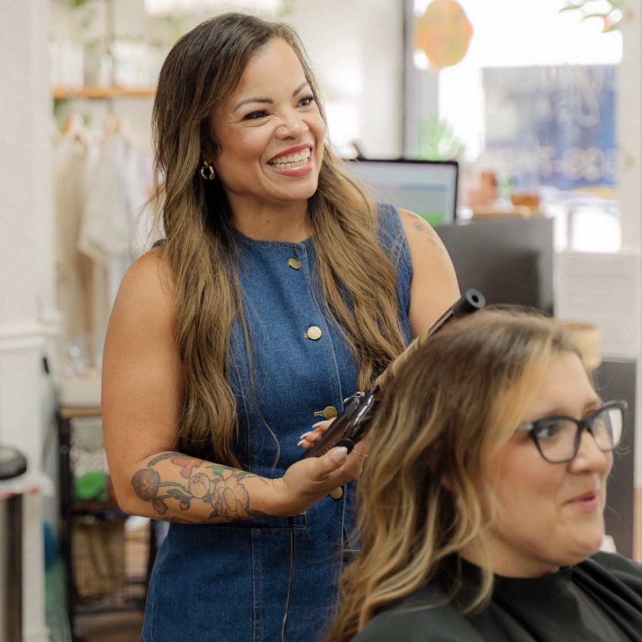 Smiling woman with long hair and tattoos, wearing a sleeveless denim top, holding a hairbrush in a salon.