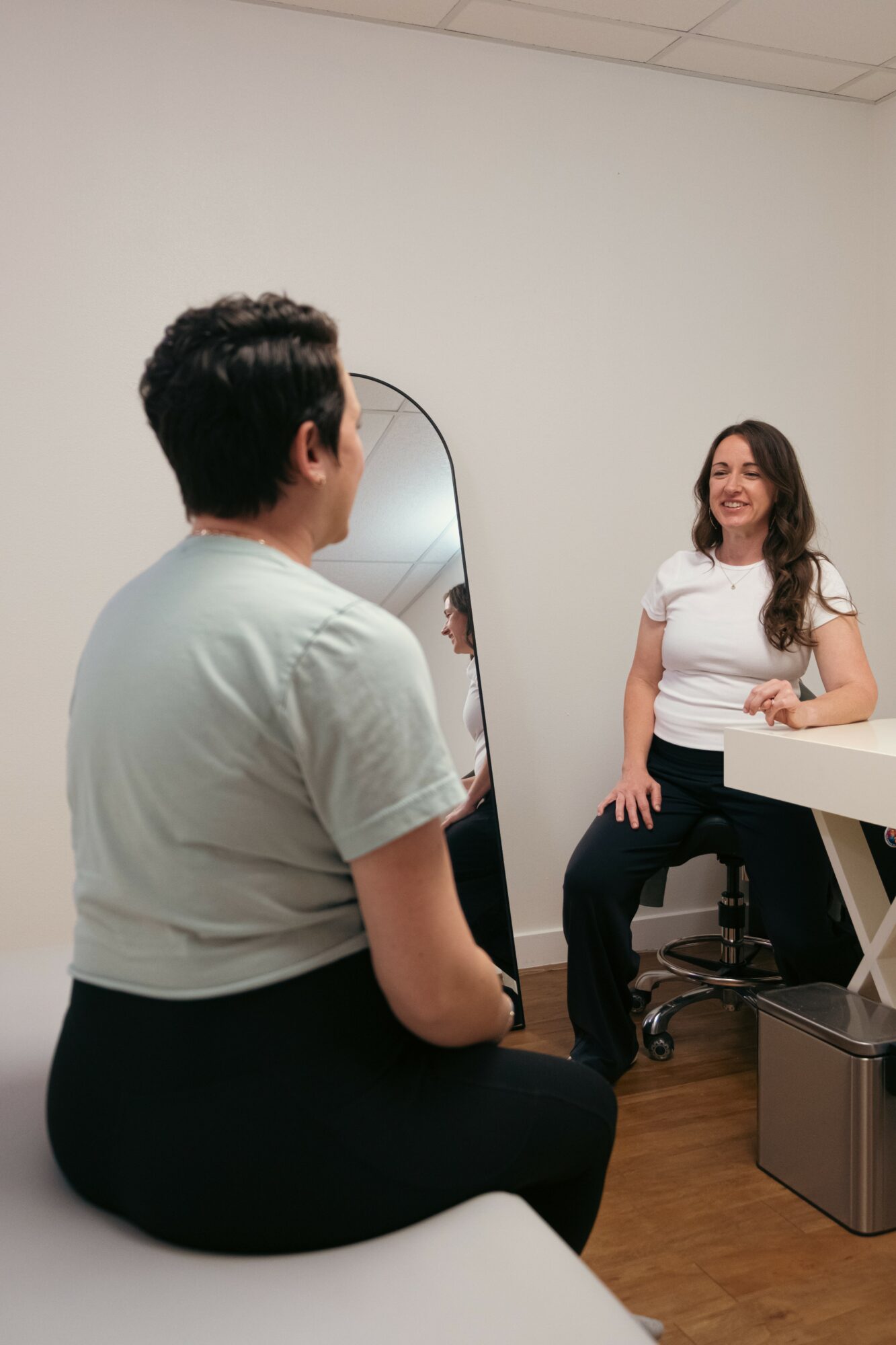 Two women sitting and talking in a room with a mirror and a desk, one smiling and the other listening.