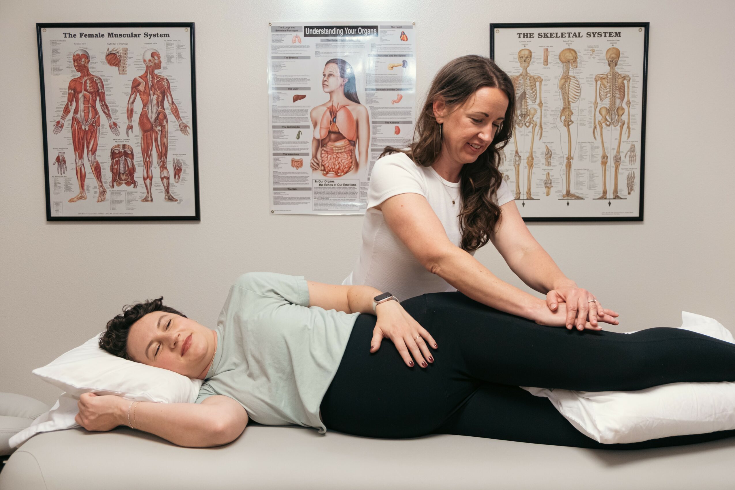 Woman lying on bed receiving a massage from a therapist in a room with anatomical posters on the wall.