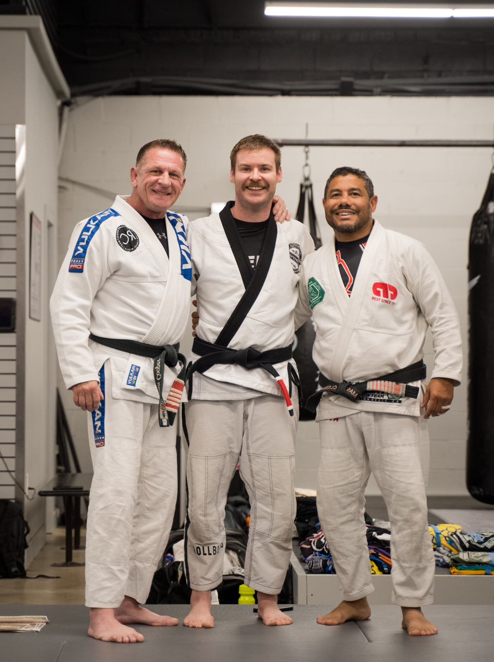 Three men in Brazilian Jiu-Jitsu gis standing together indoors, smiling, with a white wall and equipment in background.