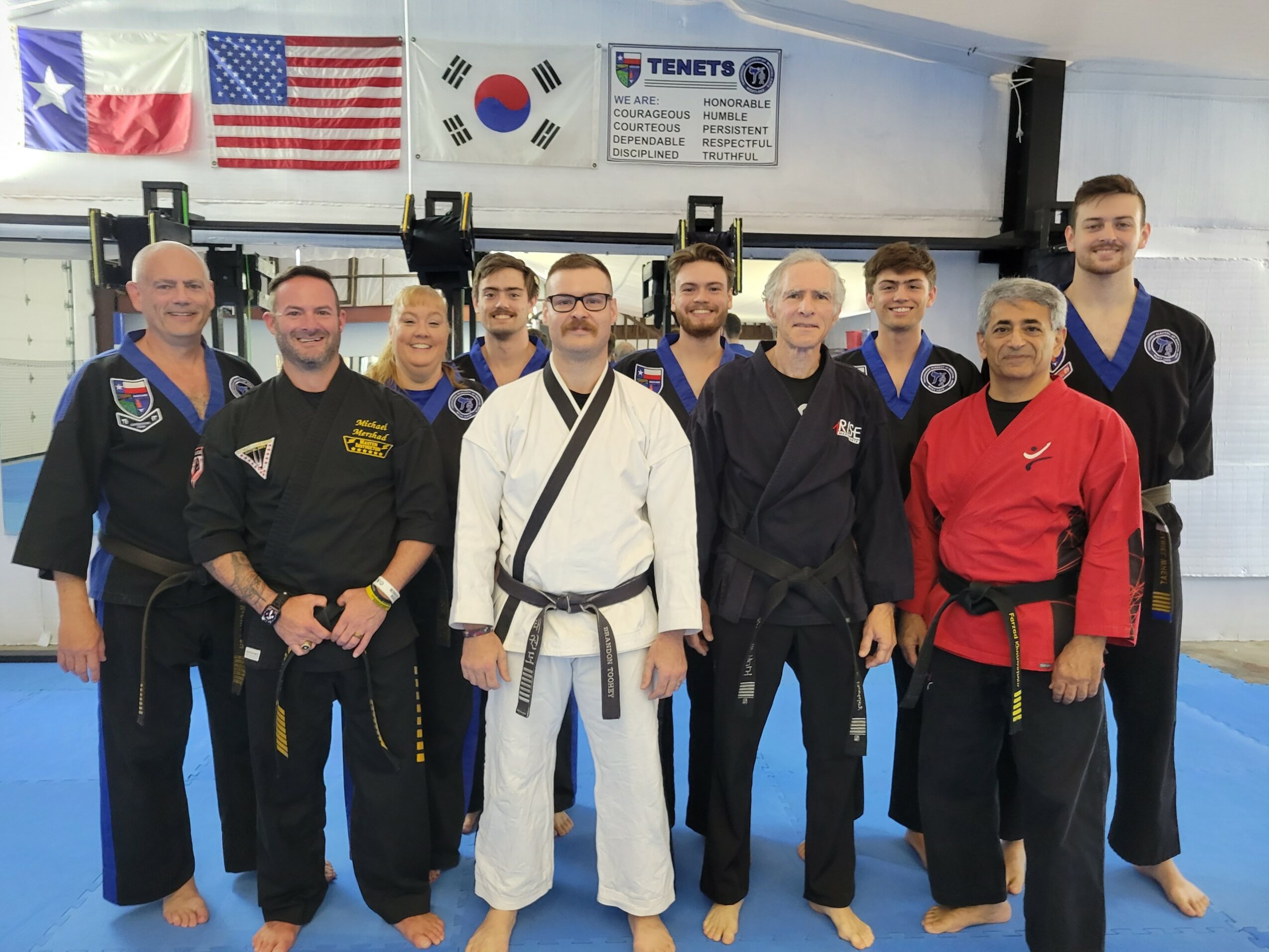 Group of martial artists in uniform, standing in dojo with flags and banners, smiling for photo.