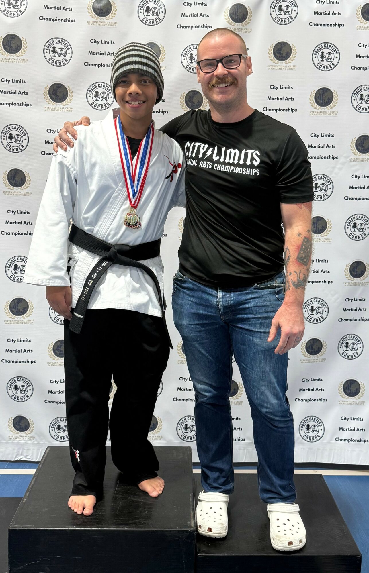 Two people standing on a podium, one wearing a martial arts uniform with a medal, the other in casual clothing, smiling.