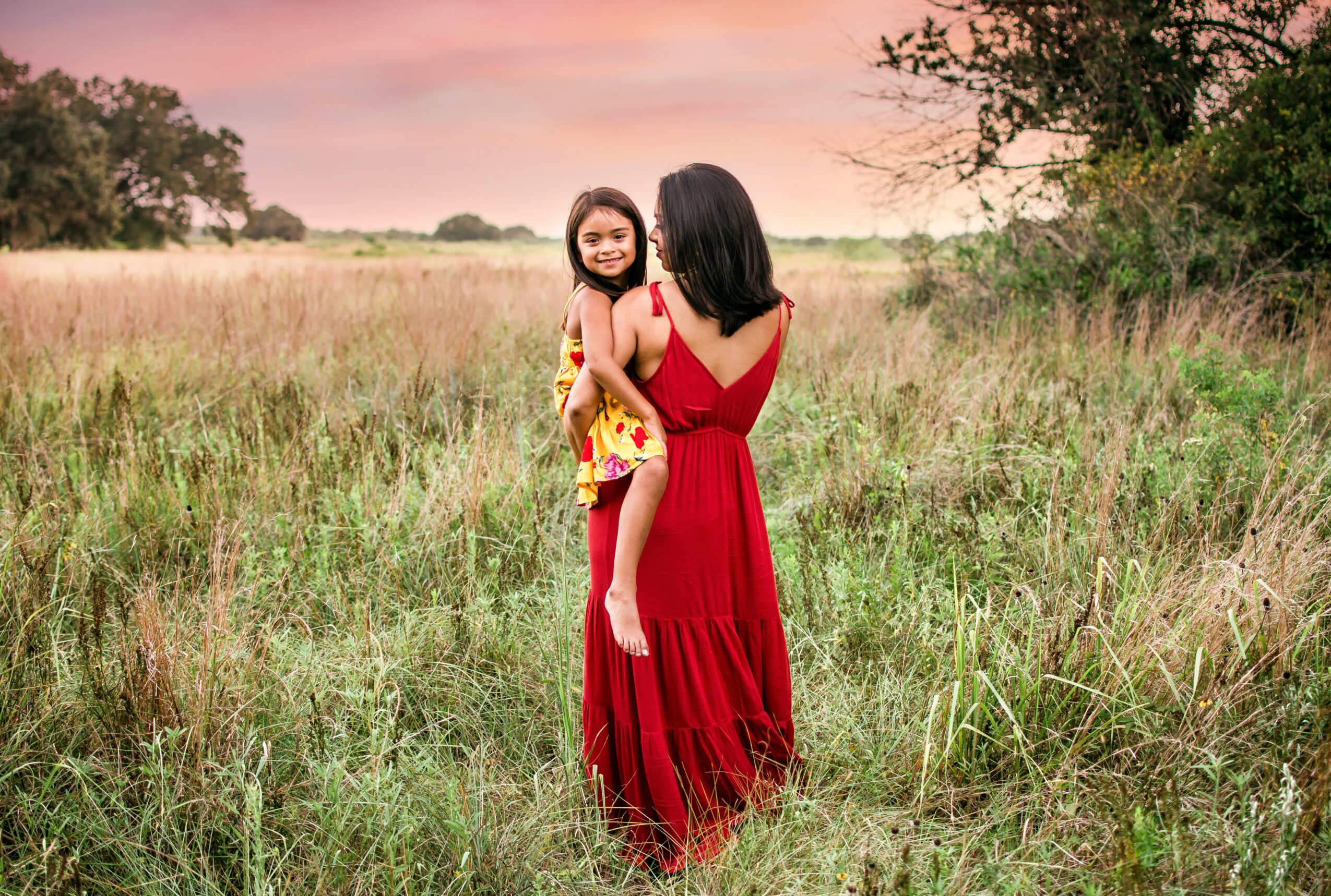Two women standing in a grassy field at sunset, one holding a child, all smiling, with trees in the background.