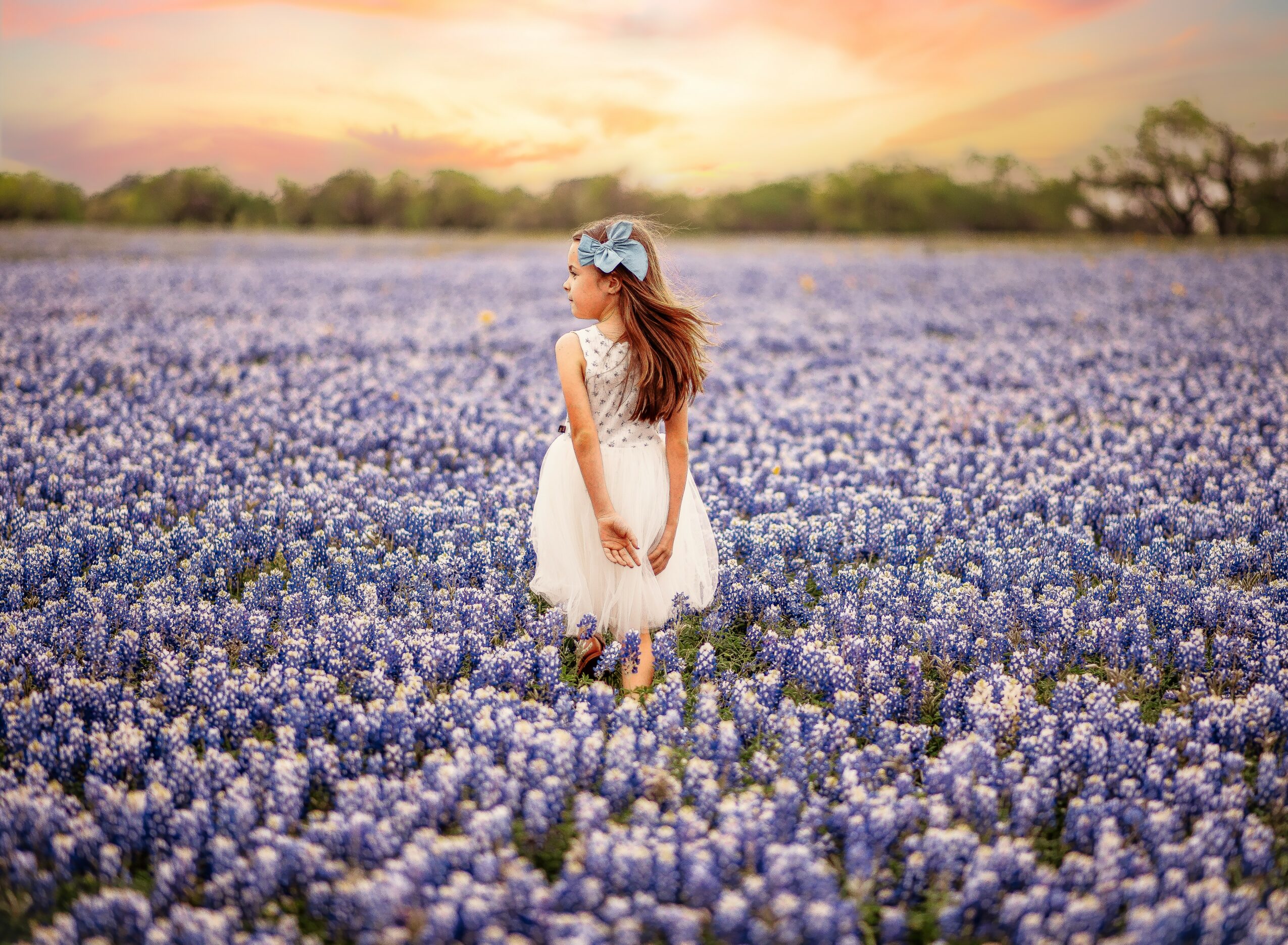 Girl in a white dress and headband standing in a purple flower field at sunset, facing sideways.