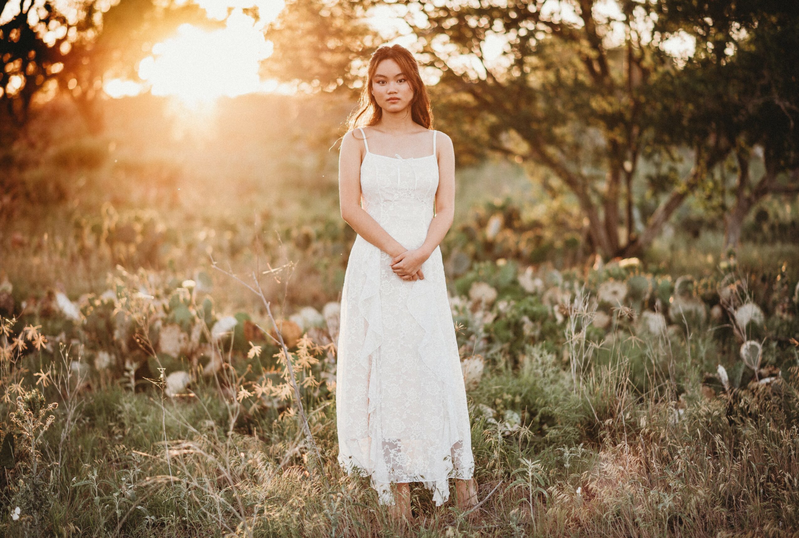 Young woman in a white dress standing in a field with trees and sunlight in the background.