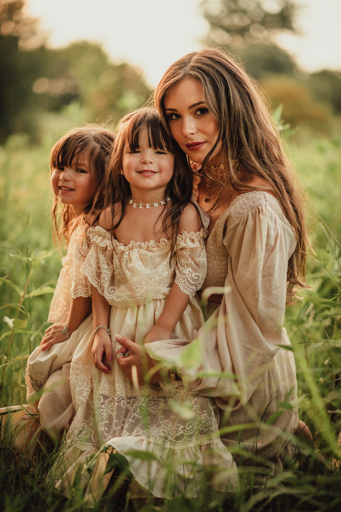 Three females sitting outdoors in a grassy field, smiling, with trees in the background, wearing light-colored dresses.