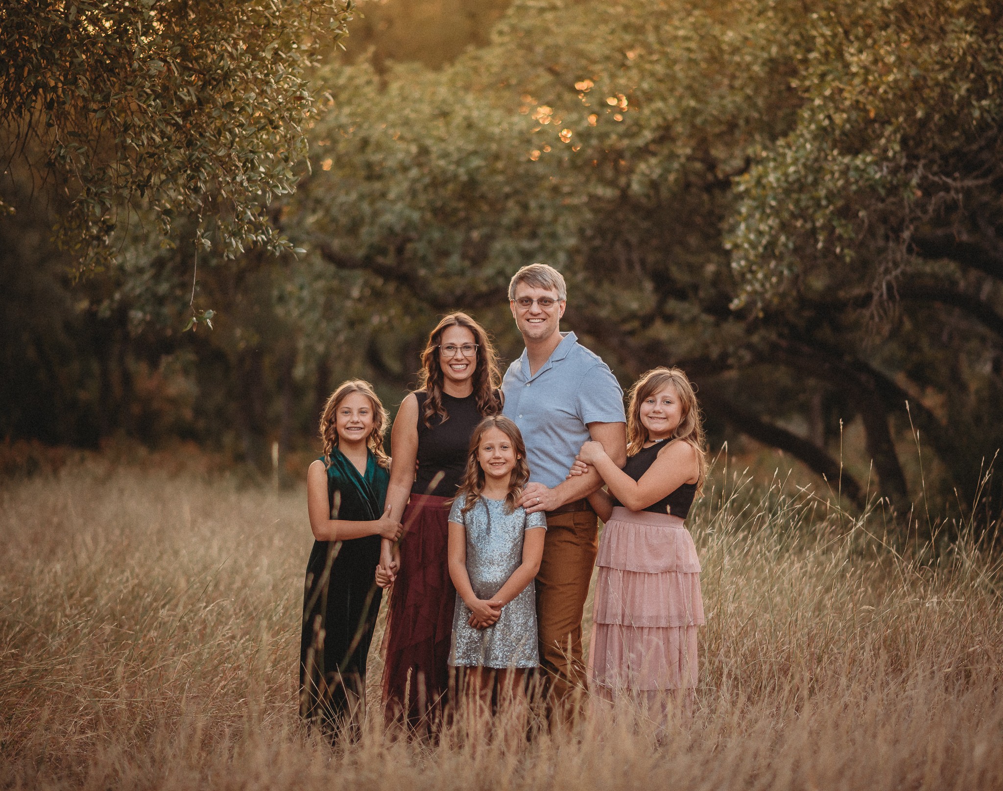 Family of five standing in a grassy field with trees in the background, smiling at the camera.