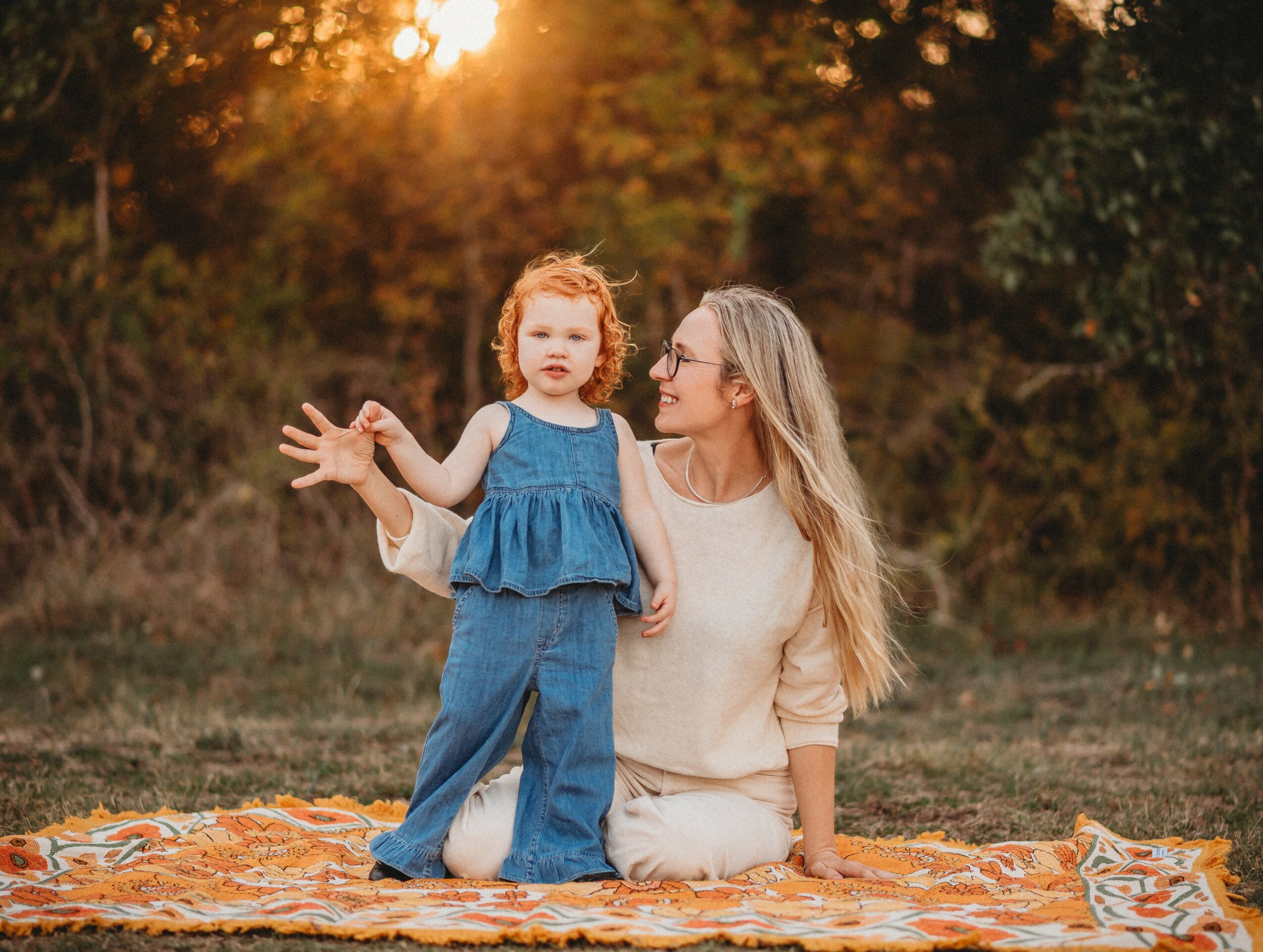 Woman and young girl sitting on a blanket outdoors during sunset, girl gesturing with hand, woman looking at girl.