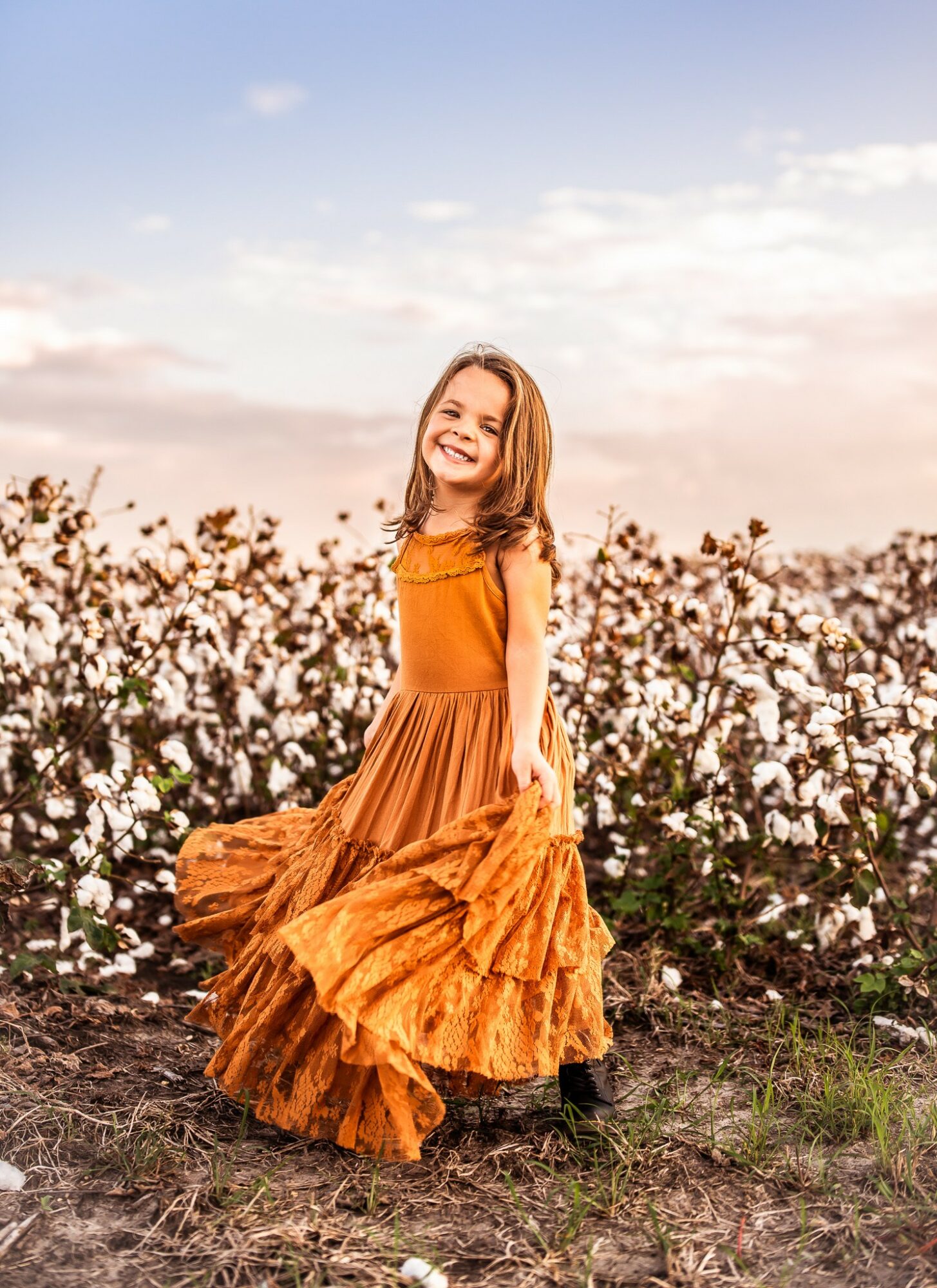 Young woman in an orange dress standing outdoors among flowering bushes, smiling, with a sky background.