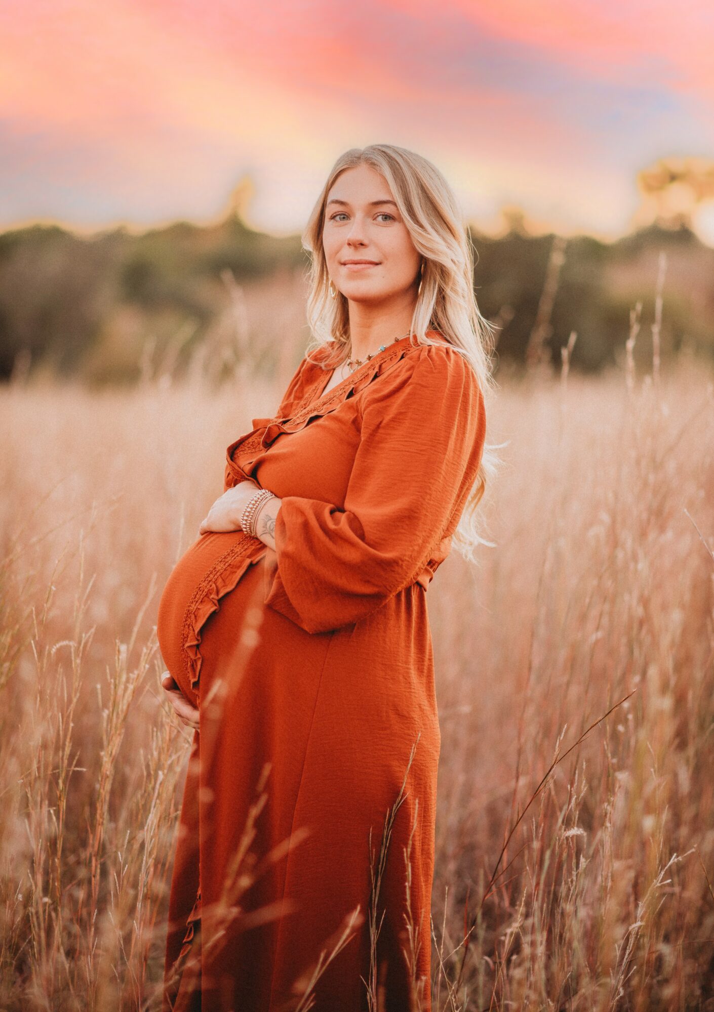 Young woman with blonde hair in a field of tall grass at sunset, wearing an orange dress, smiling softly.