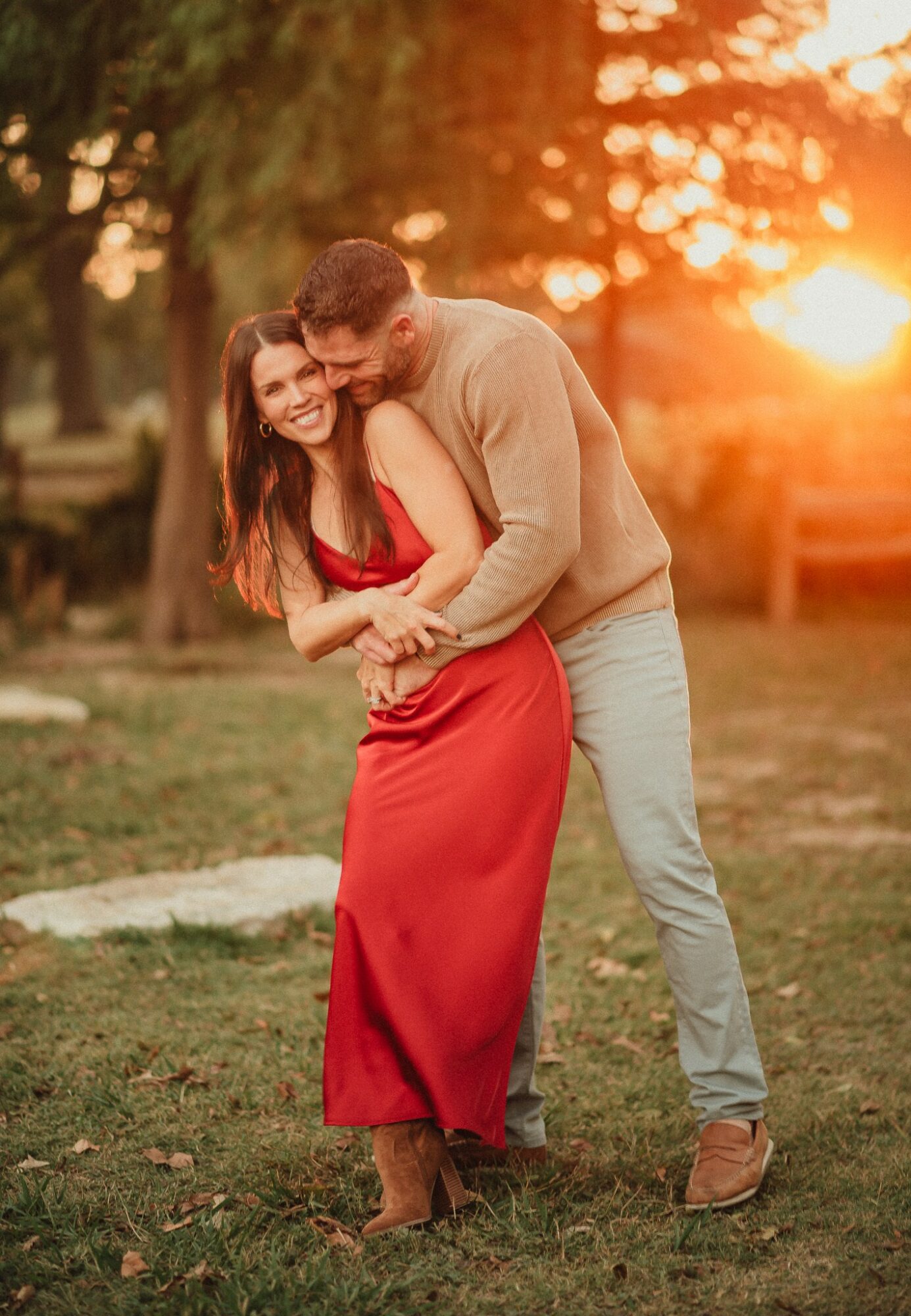 A couple hugging outdoors during sunset, woman in a red dress, man in a beige sweater and light pants, smiling.