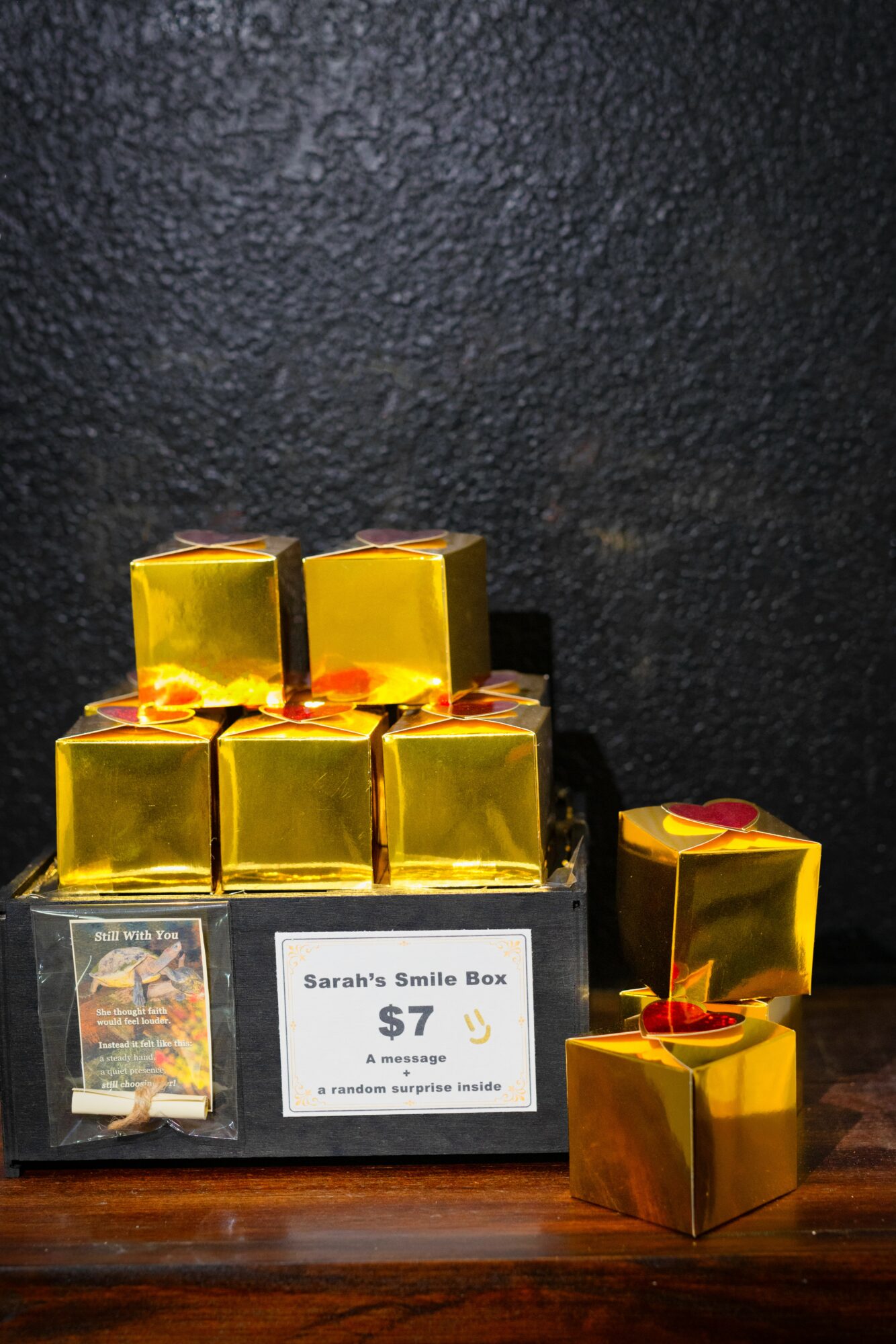 Small yellow boxes with red ribbons arranged on a wooden surface against a dark background, labeled as Sarah's Smile Box, priced at $7.