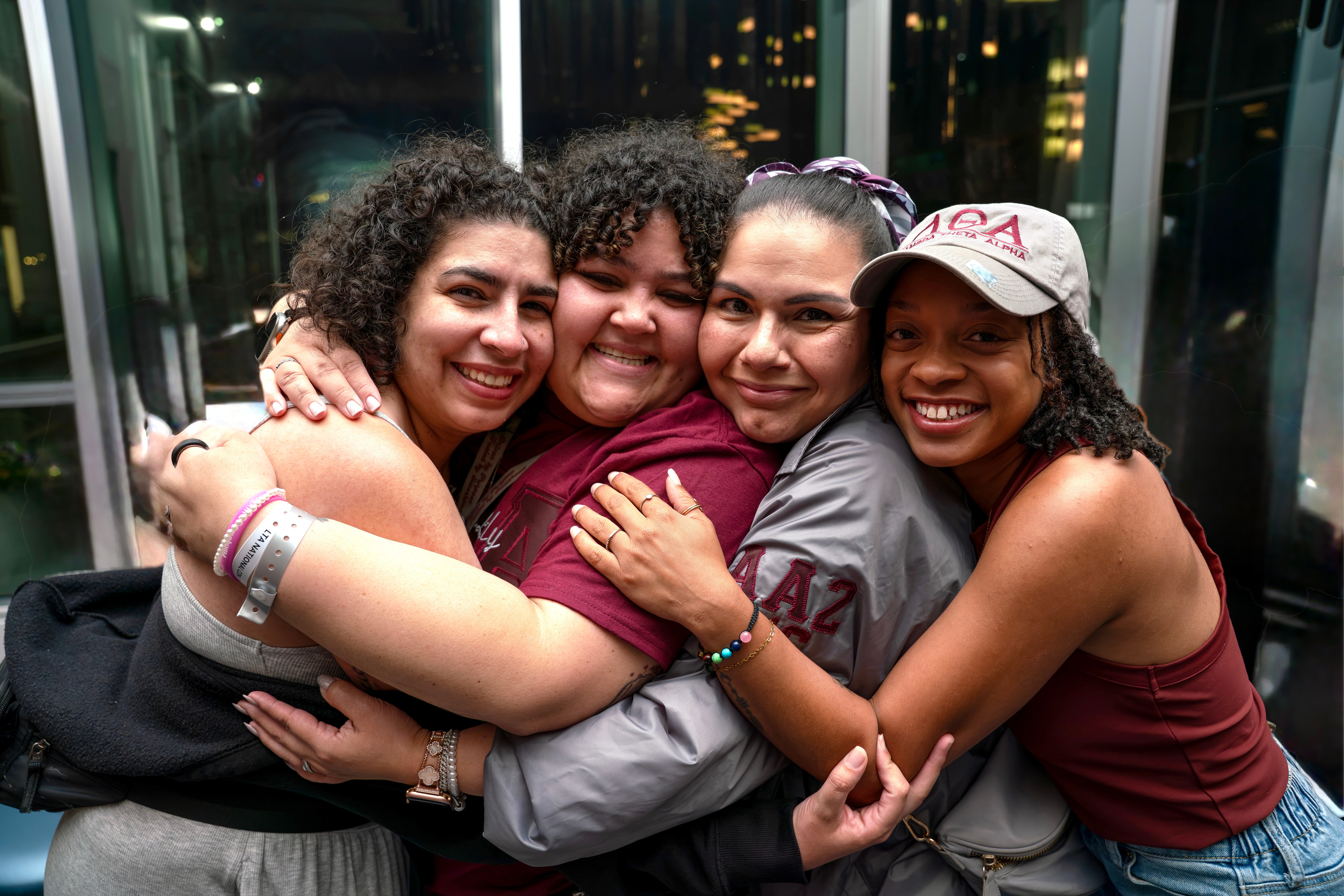 Four women hugging and smiling indoors with glass windows in the background.
