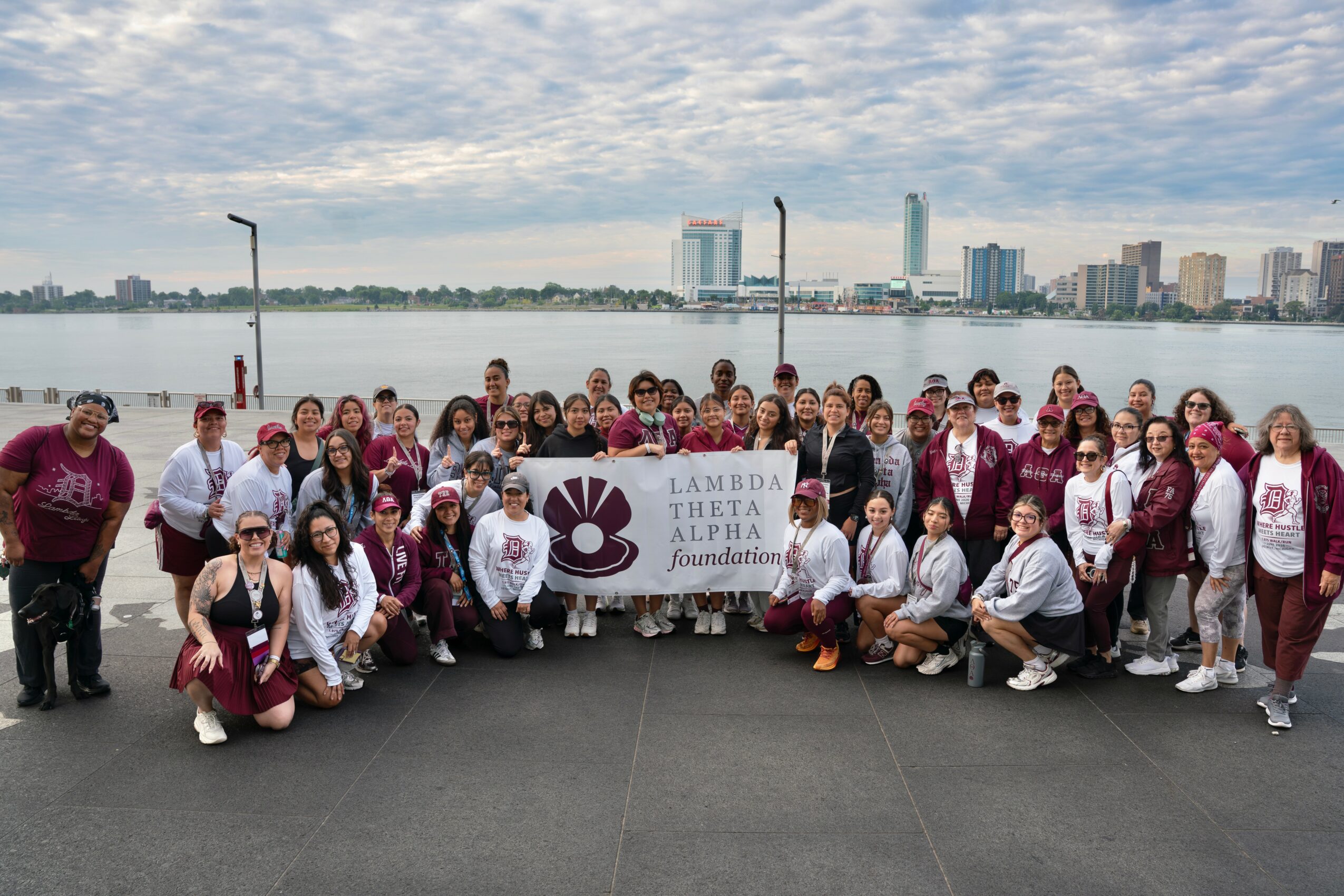 Group of people holding a Lambda Theta Phi banner by a river with city skyline in the background, overcast sky.