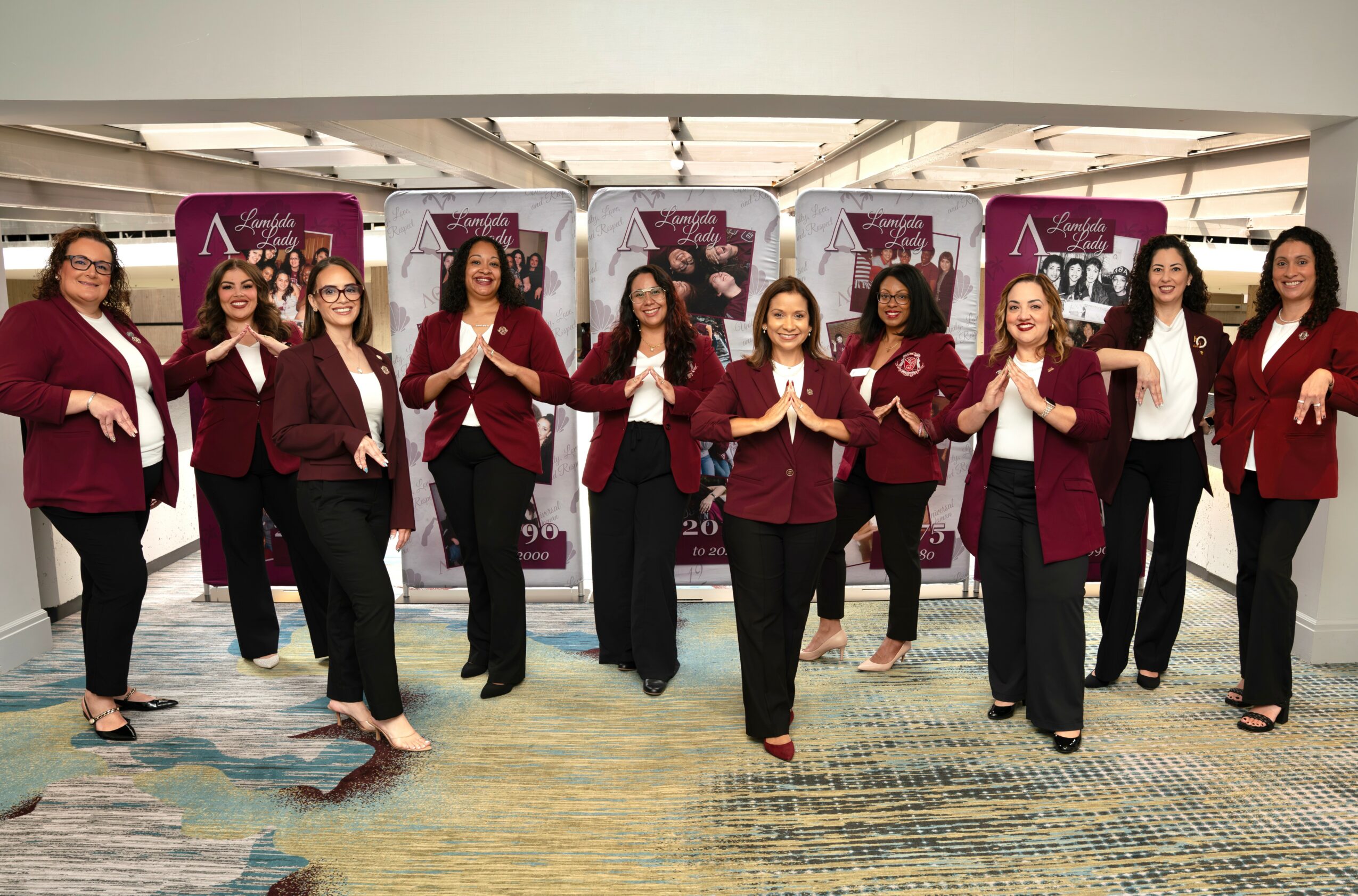 Group of ten women in matching maroon blazers and black pants standing in a line indoors, smiling and holding hands.