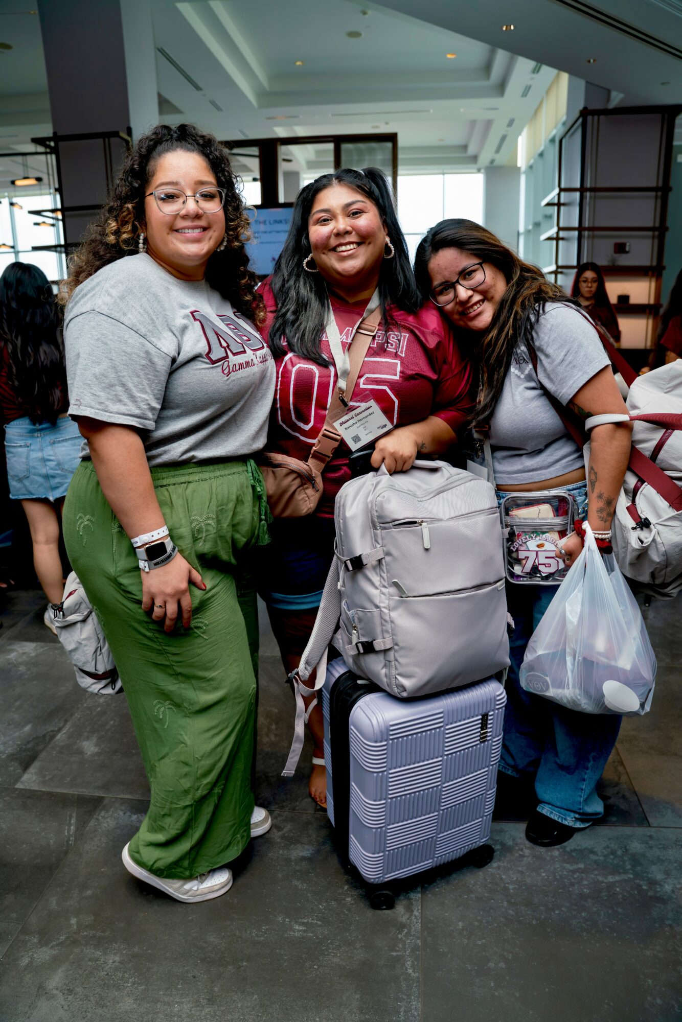 Three smiling women with backpacks and luggage in an indoor setting, standing close together, facing the camera.