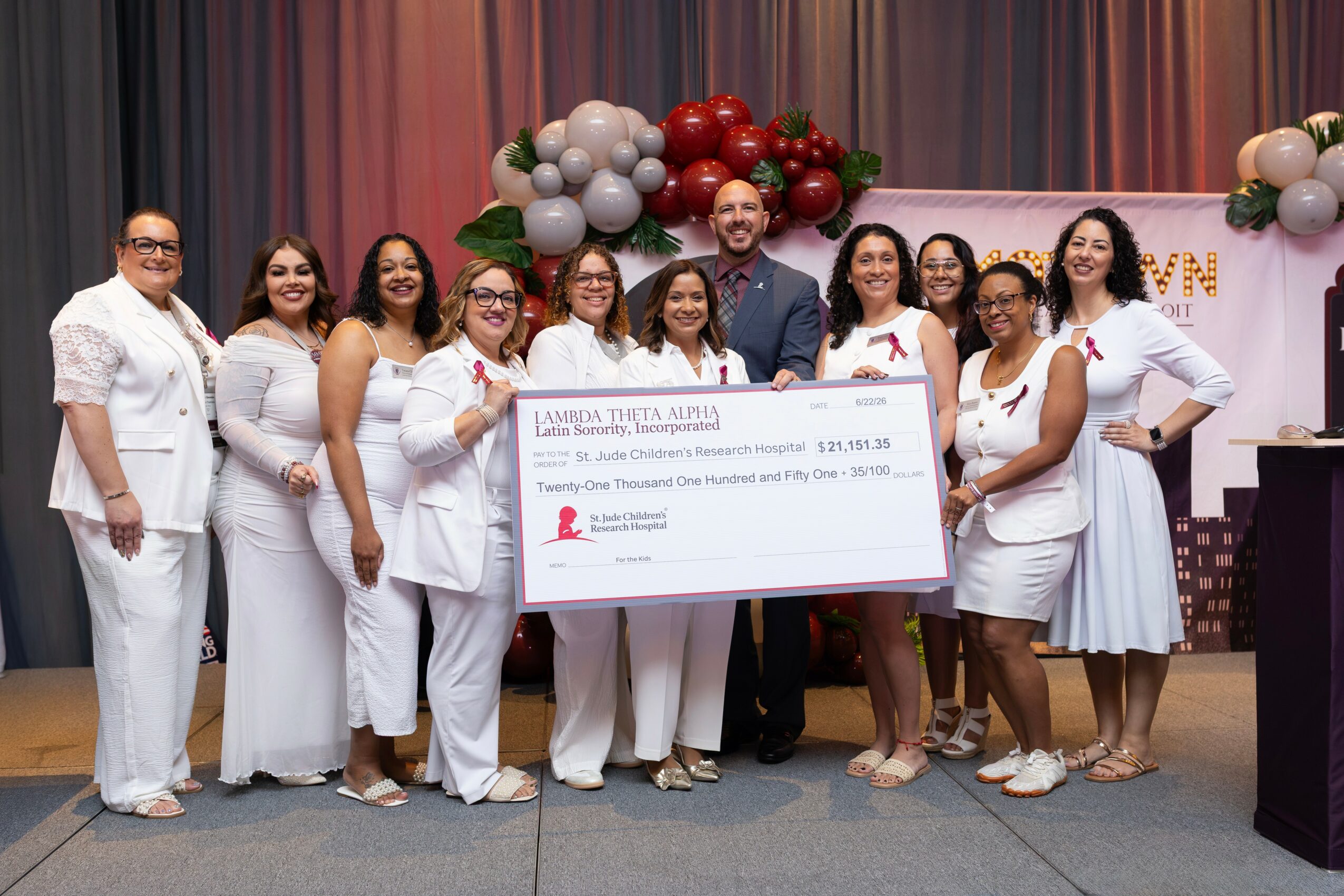 Group of women in white outfits holding a large check on stage with colorful balloon decorations behind them.