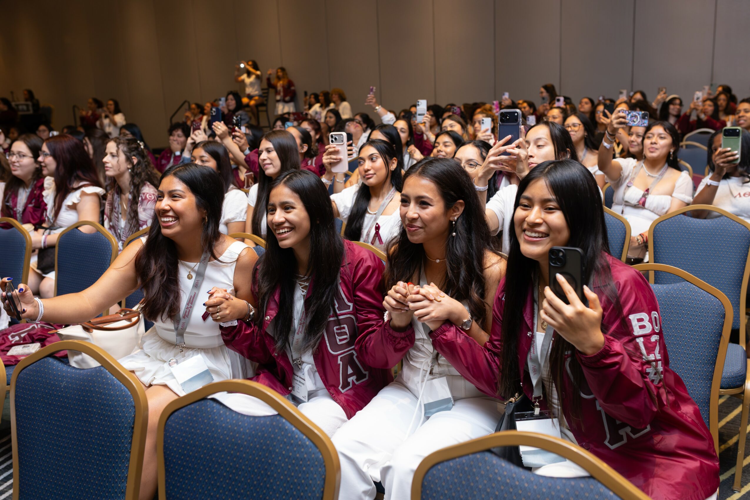 Group of young women sitting in an auditorium, smiling and taking photos, with many people in the background.