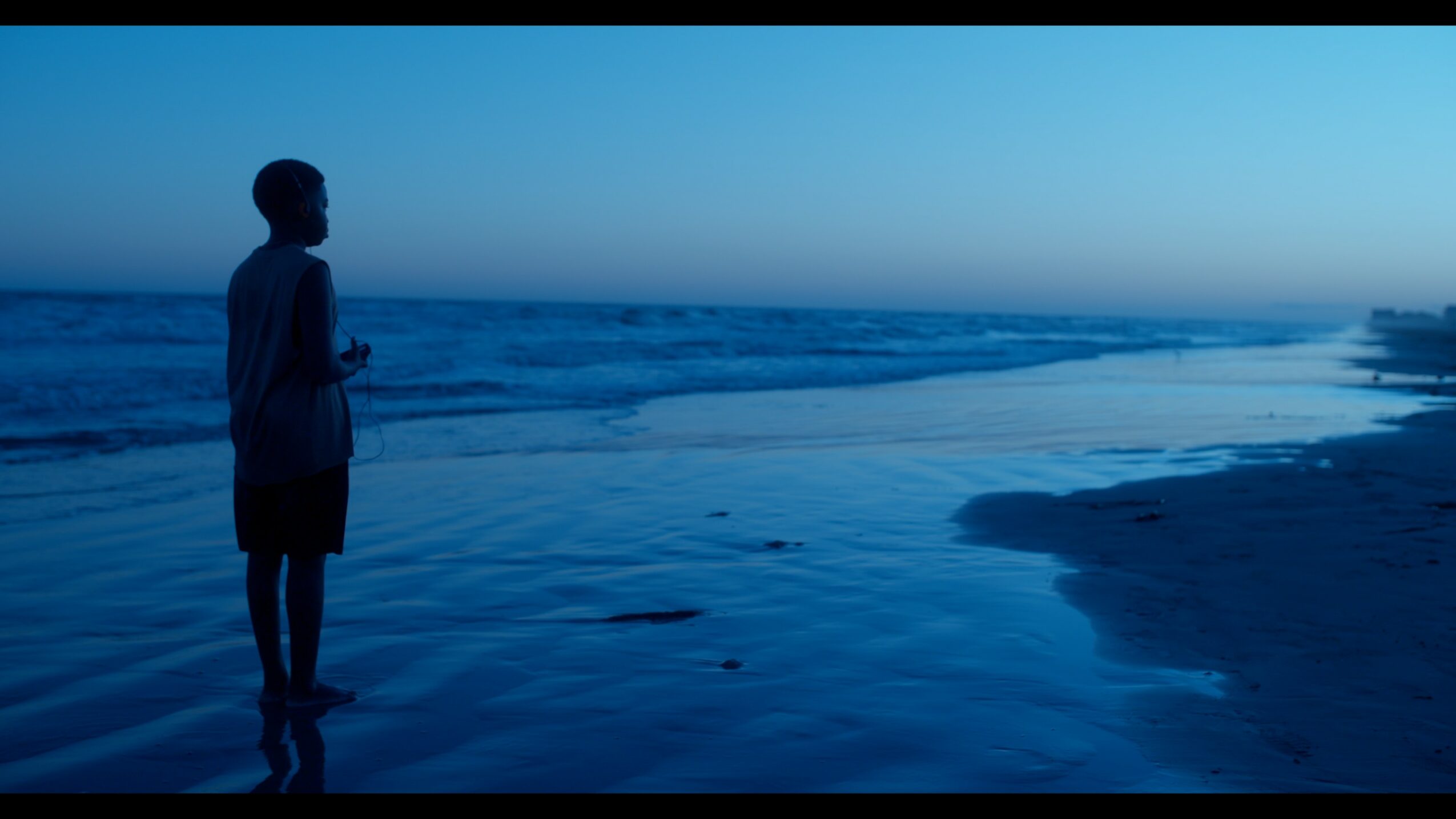 Person standing on the beach near the water, looking at the ocean during dusk or dawn.