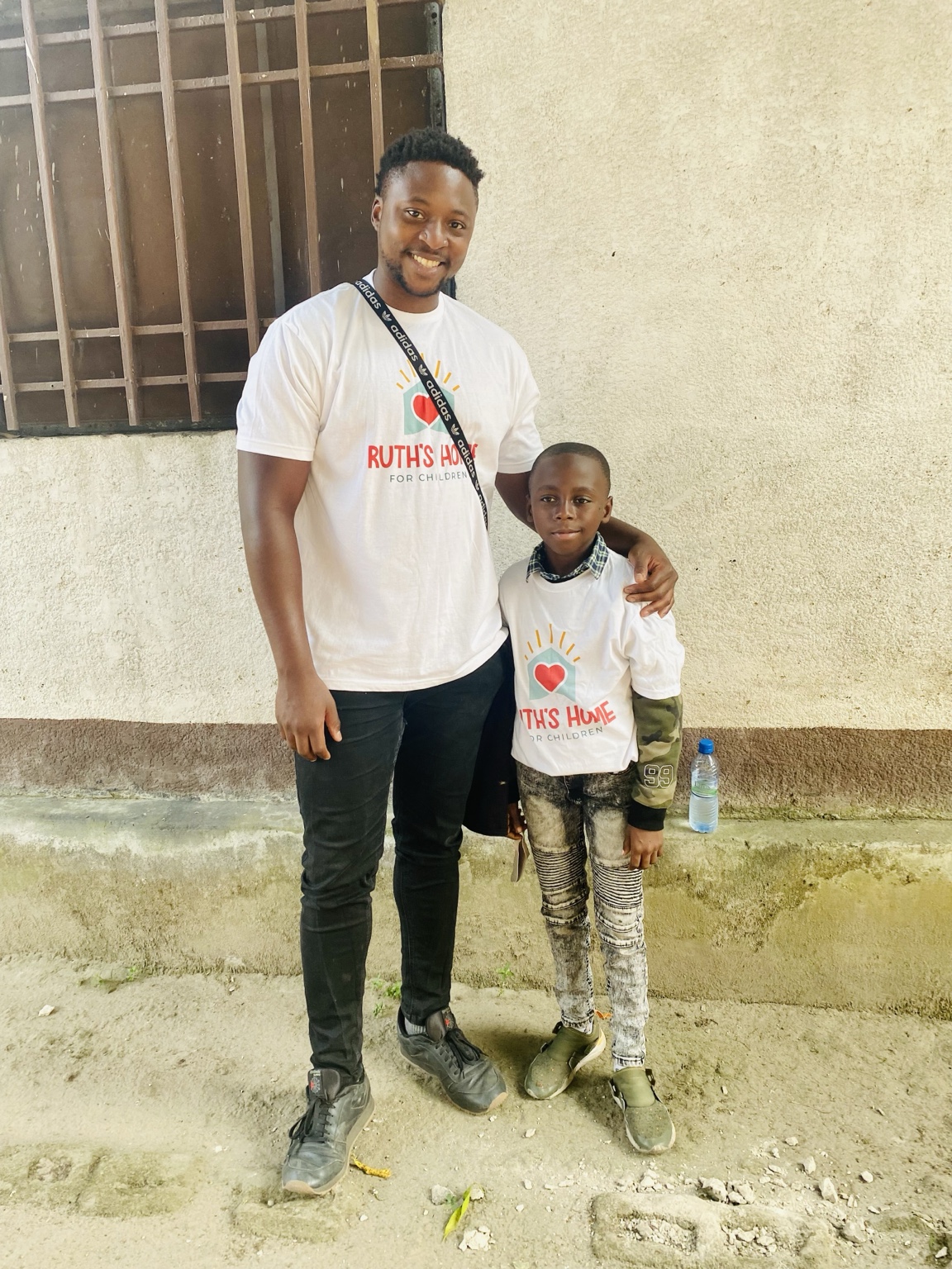 Two people, a woman and a child, stand outdoors in front of a wall with a window. Both wear white T-shirts with red text and a heart symbol. The woman has her arm around the child's shoulder.