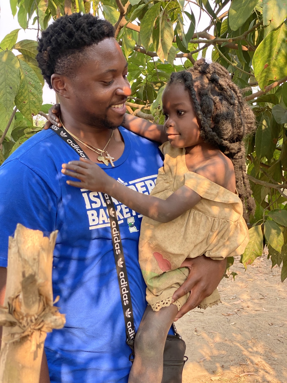 Adult holding a young girl outdoors among green leaves, both smiling and looking at each other.