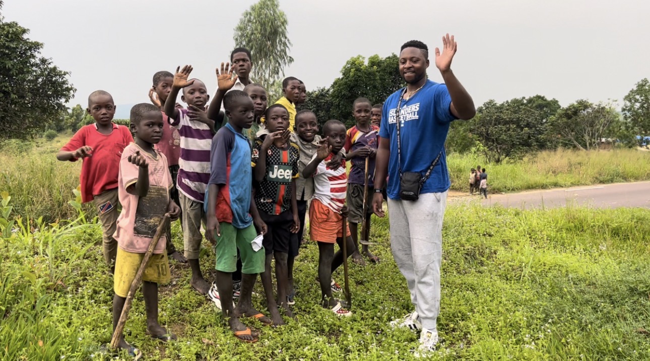 Group of children and a man waving outdoors in a grassy field, trees in the background.