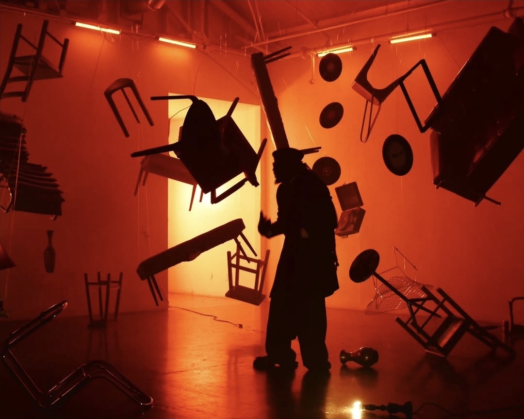 Person standing in a room with chairs and objects suspended in mid-air, illuminated by orange lighting.