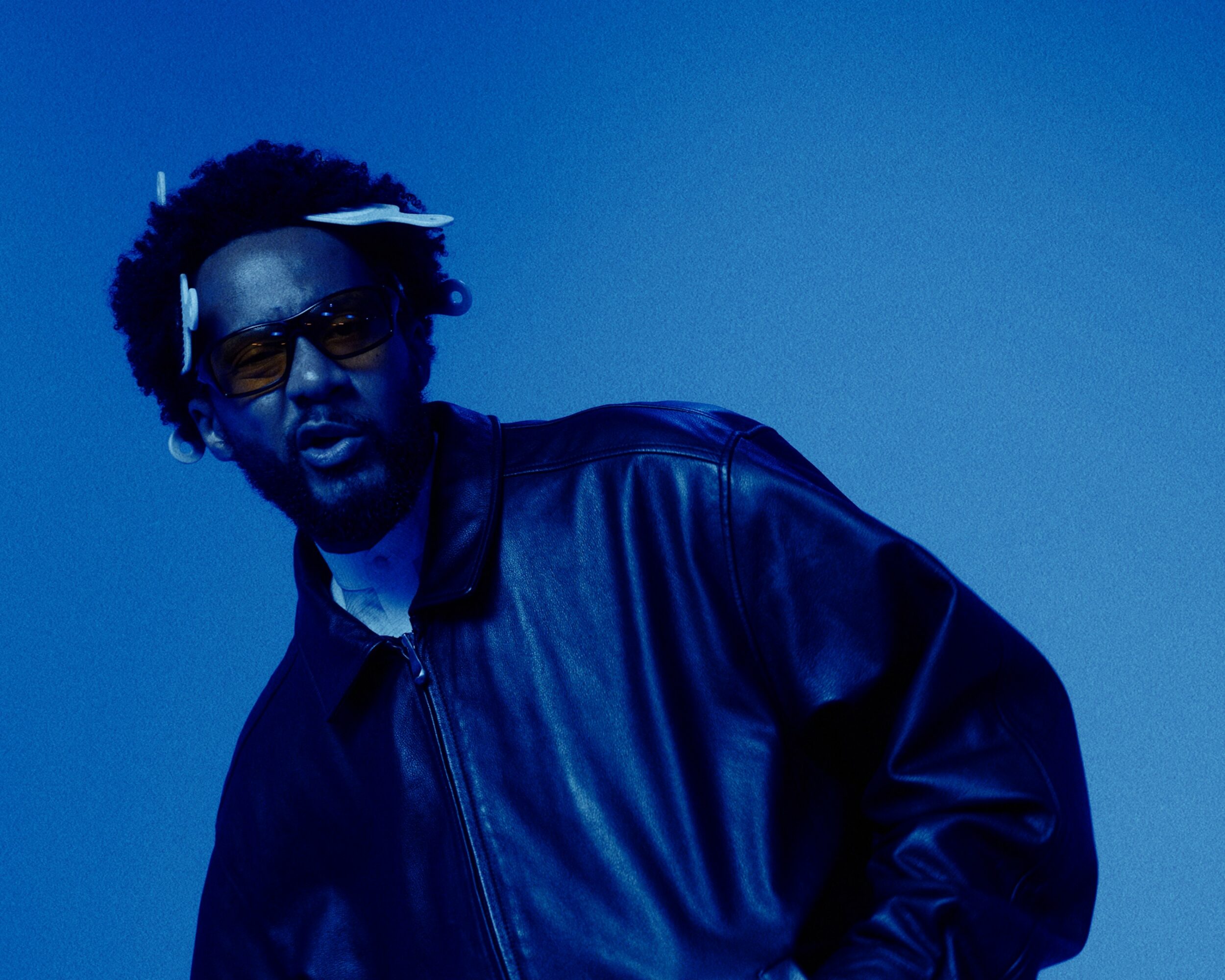 Person with sunglasses and headphones wearing a leather jacket, facing slightly to the left, against a blue background.