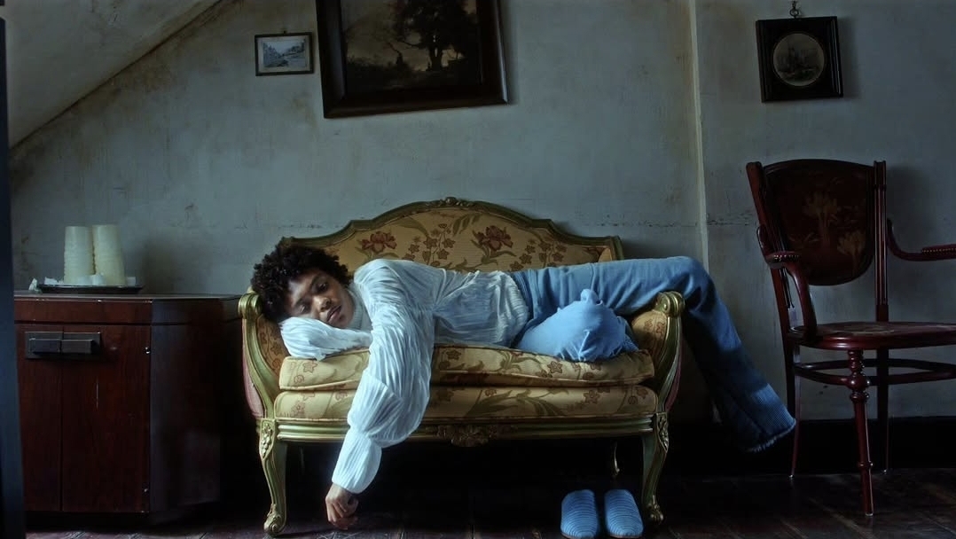 Person lying on a vintage sofa with legs hanging off, in a dimly lit room with paintings and a chair nearby.