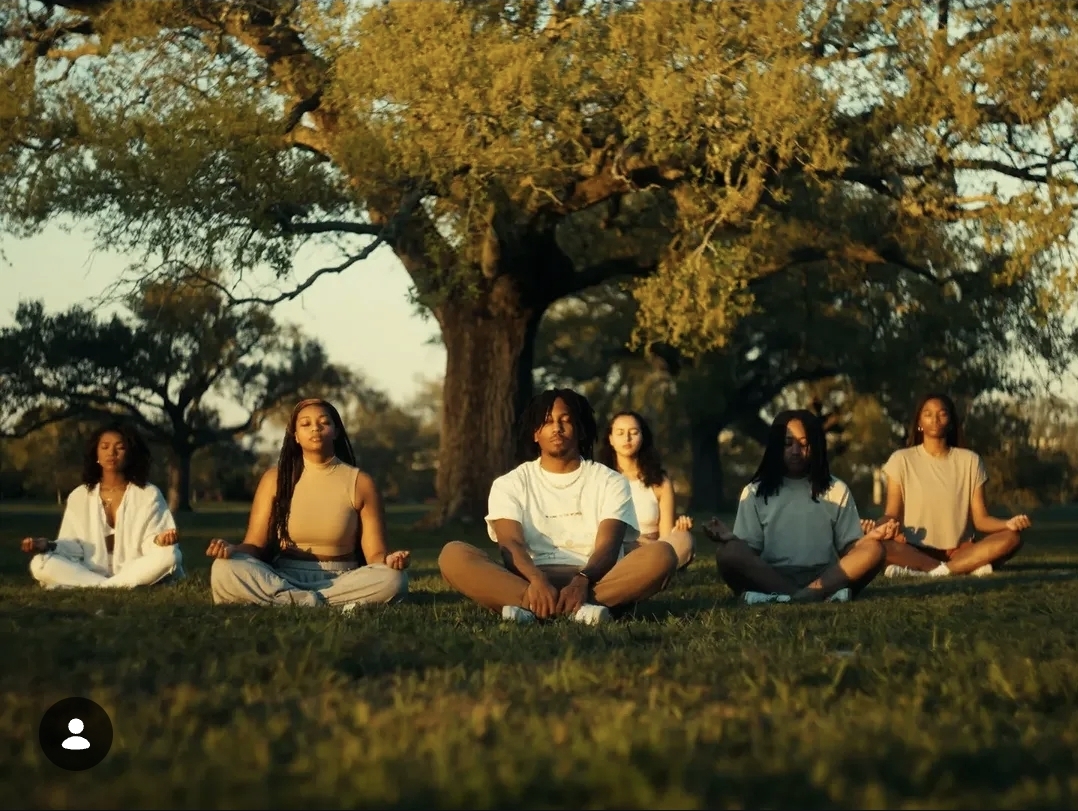 Five people sit cross-legged on grass under a large tree during sunset, practicing meditation or yoga outdoors.