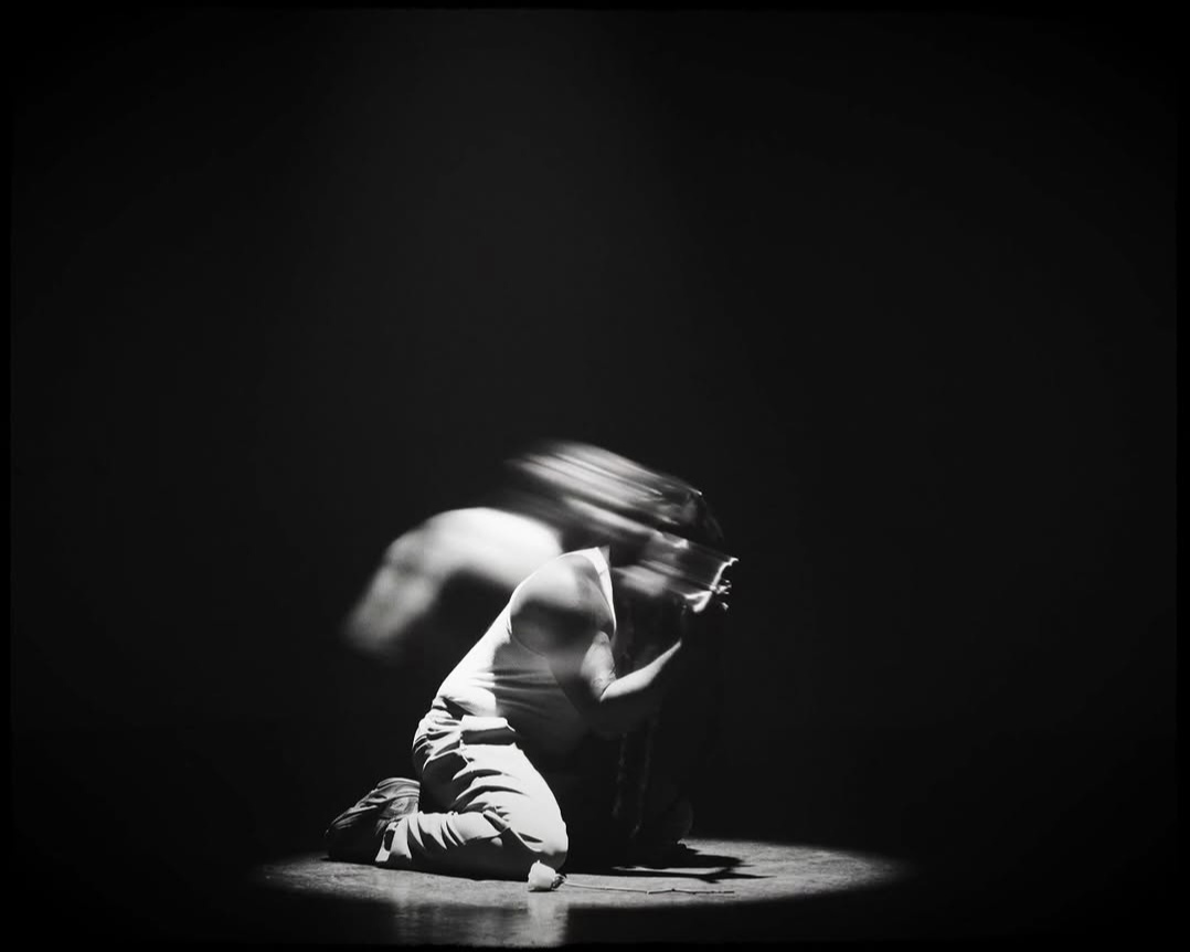 Person kneeling on the floor with head bowed and hair covering face, illuminated by a spotlight in darkness.