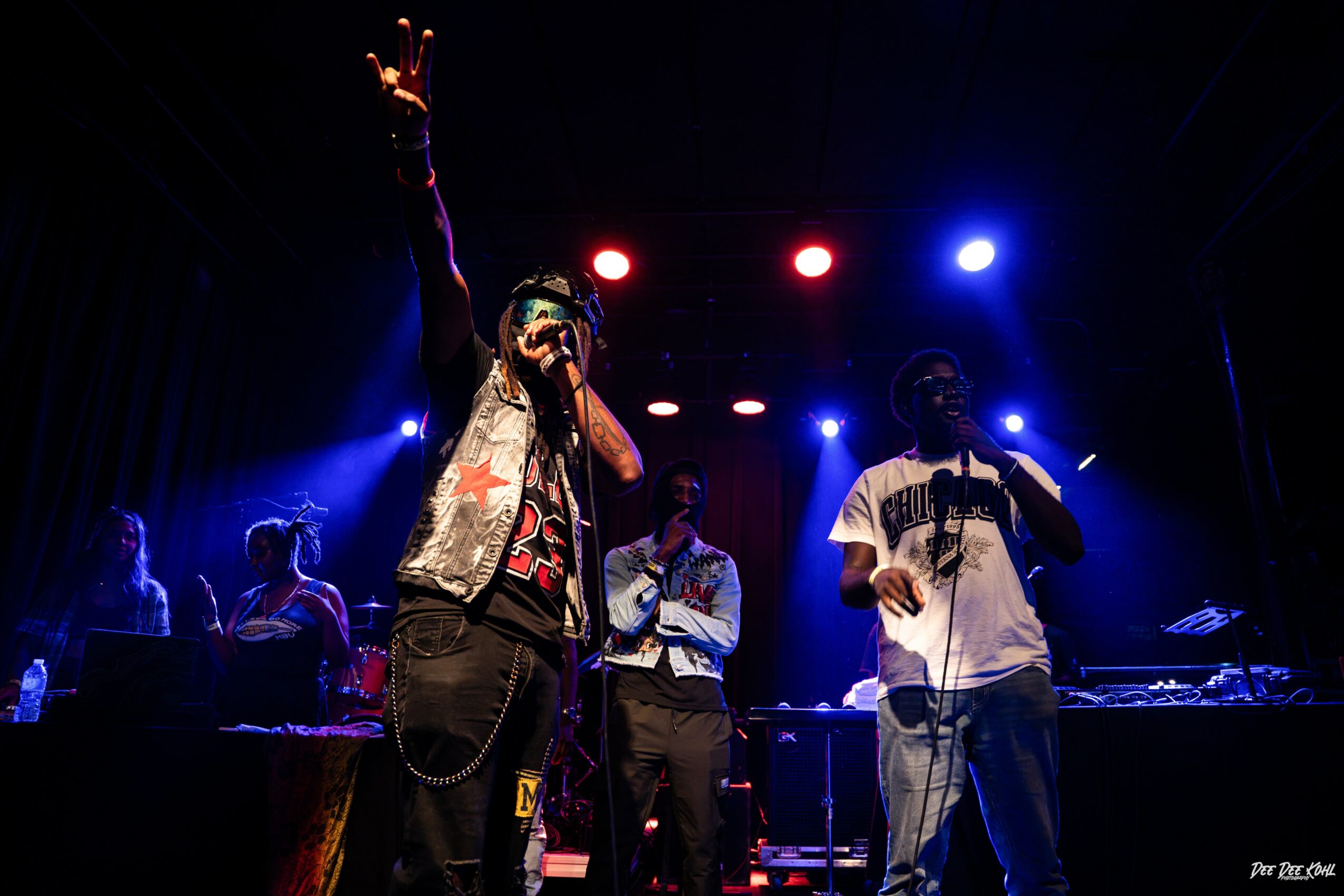 Performers on stage with colorful lights, one raising hand, others singing or DJing, dark background, lively atmosphere.